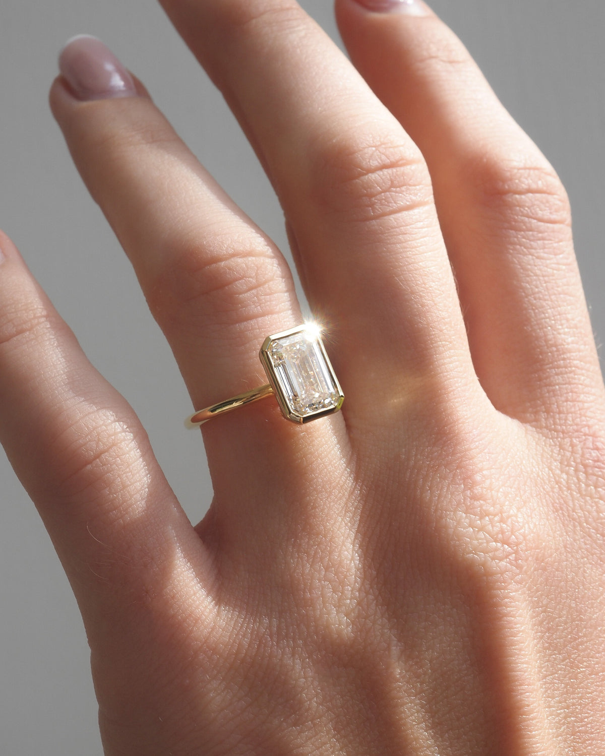 A gold ring with a emerald-cut clear diamond, worn on a person's finger against a neutral gray background.