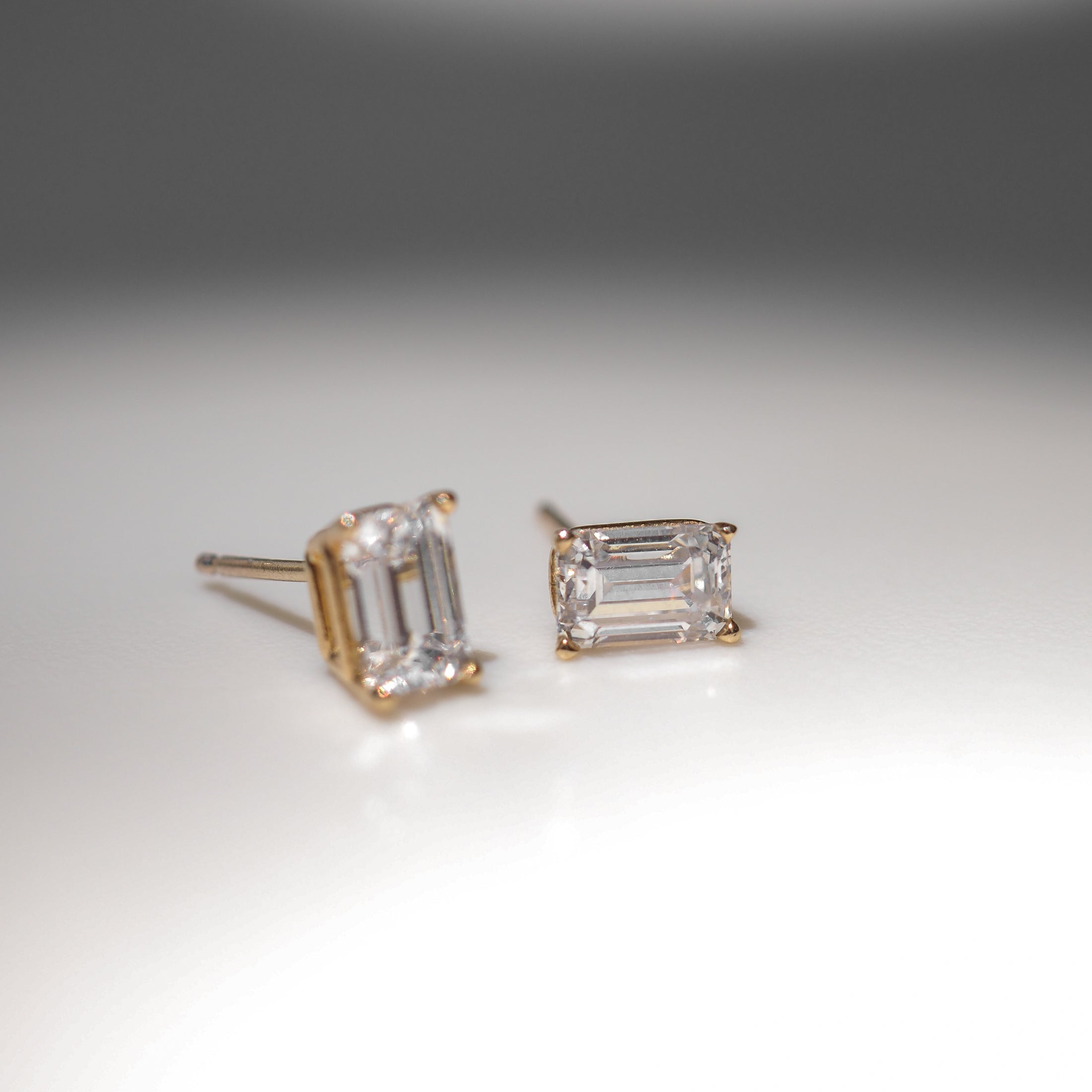 Two emerald-cut diamond stud earrings set in gold, placed on a smooth surface, showcasing high-end luxury and sophisticated jewellery design.