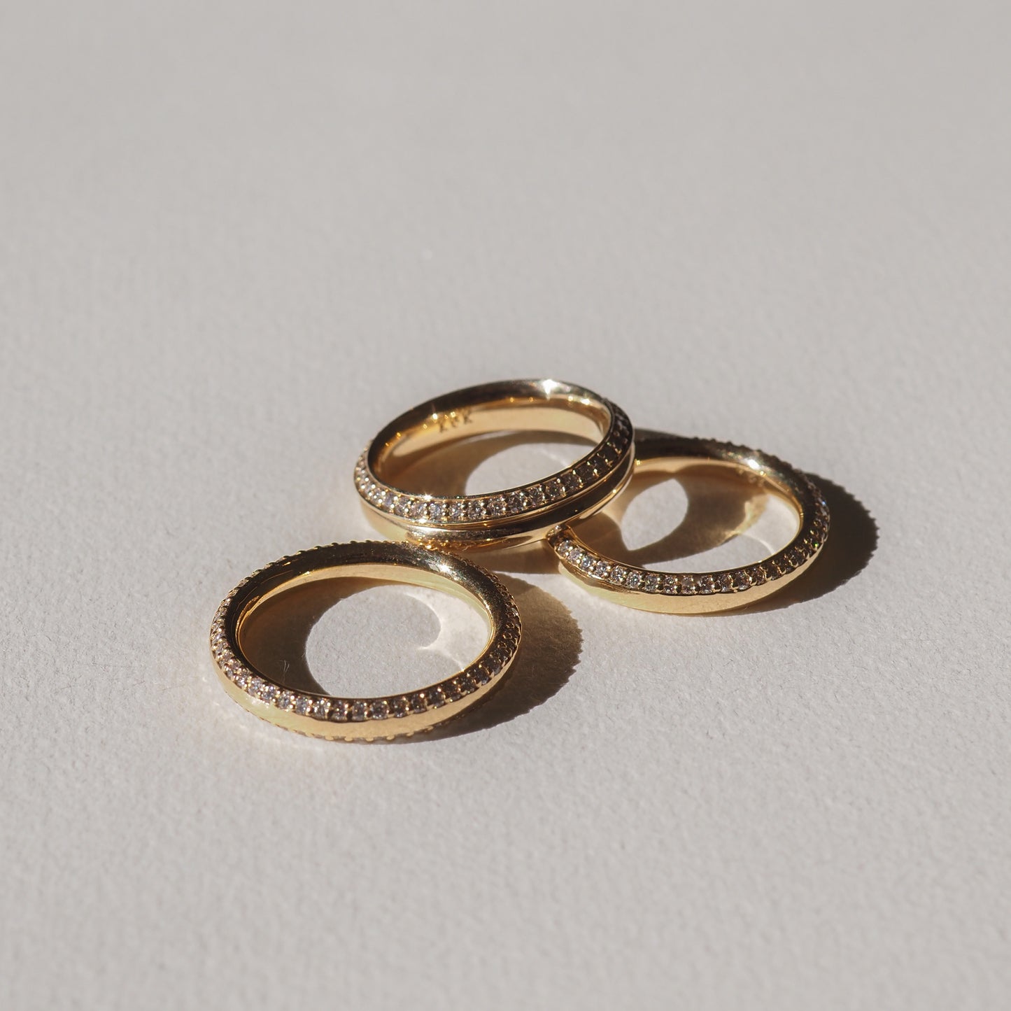 Three gold rings with diamond pavé detailing, elegantly arranged on a light surface, casting soft shadows. High-end luxury jewellery.
