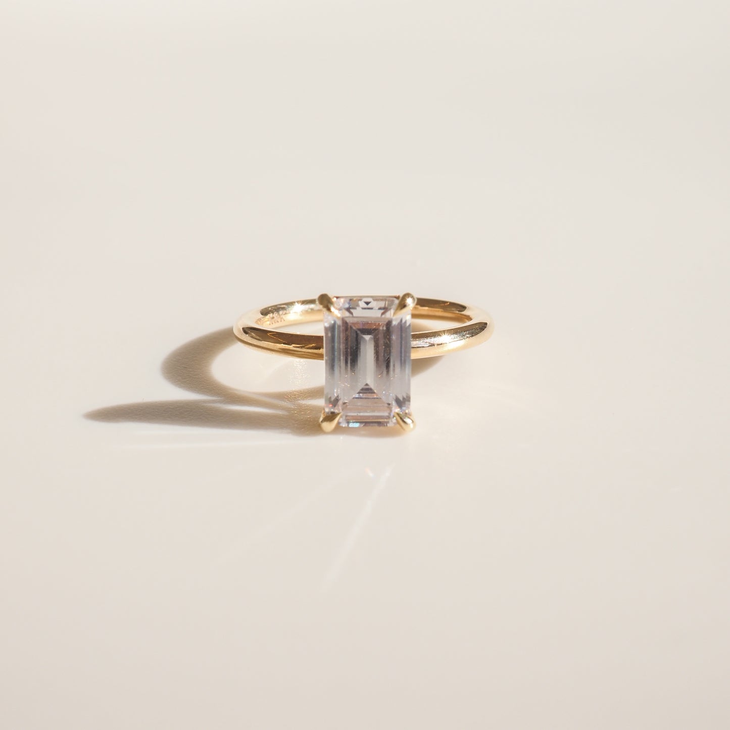 Gold ring with an emerald-cut clear gemstone on a smooth cream background, casting a delicate shadow. Perfect for luxury jewellery enthusiasts.