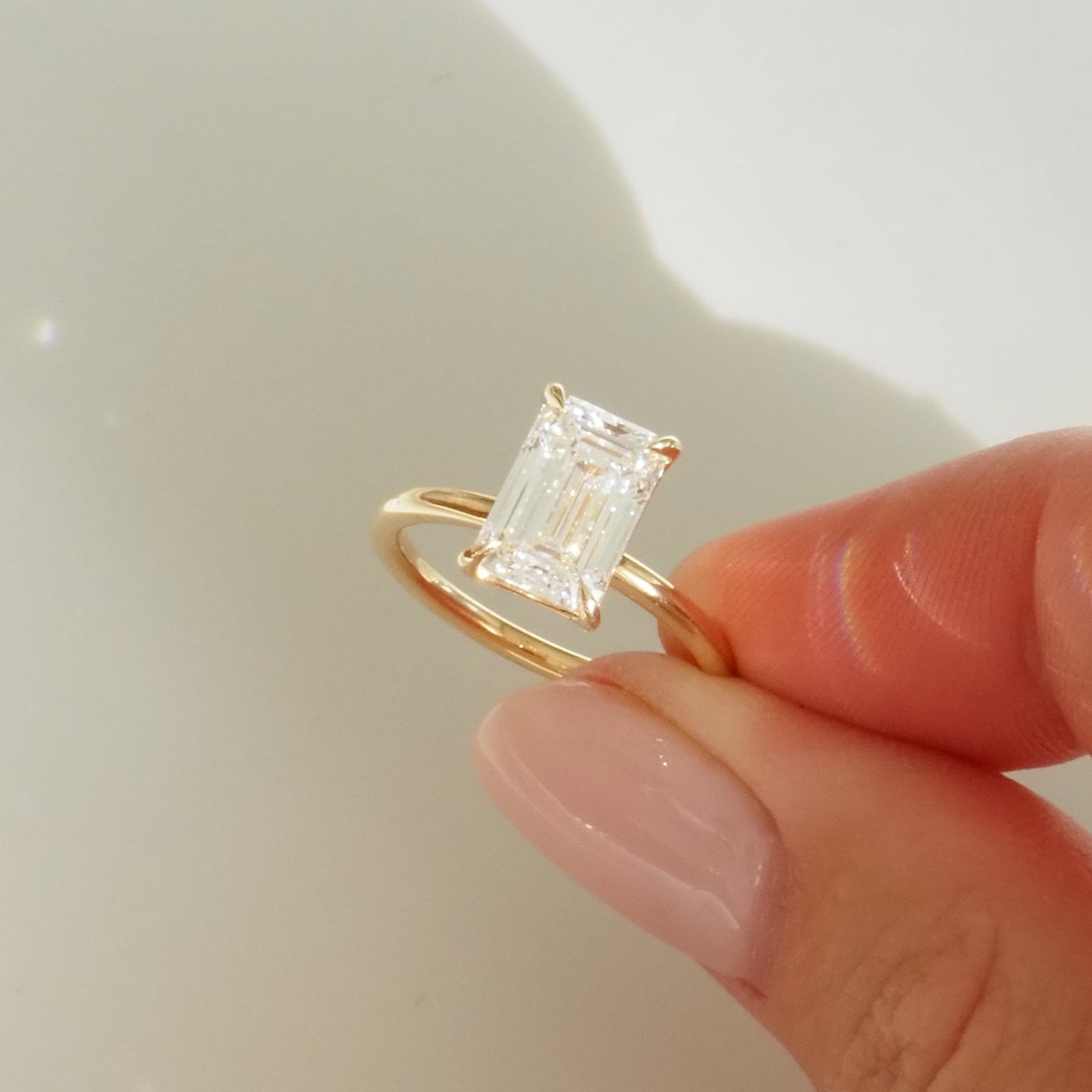 Hand holding an emerald-cut diamond ring on a gold band, reflecting light. Luxurious fine jewellery, elegant and high-end design.