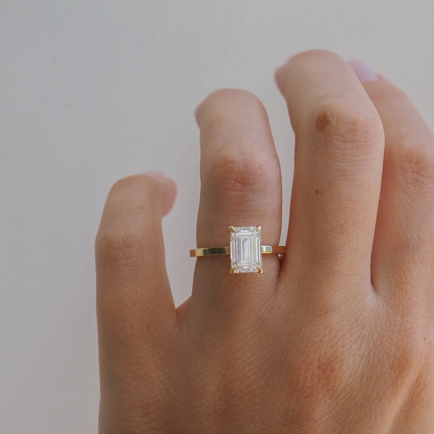 Hand wearing an emerald-cut diamond ring with a gold band on a neutral background; luxury jewellery detail.