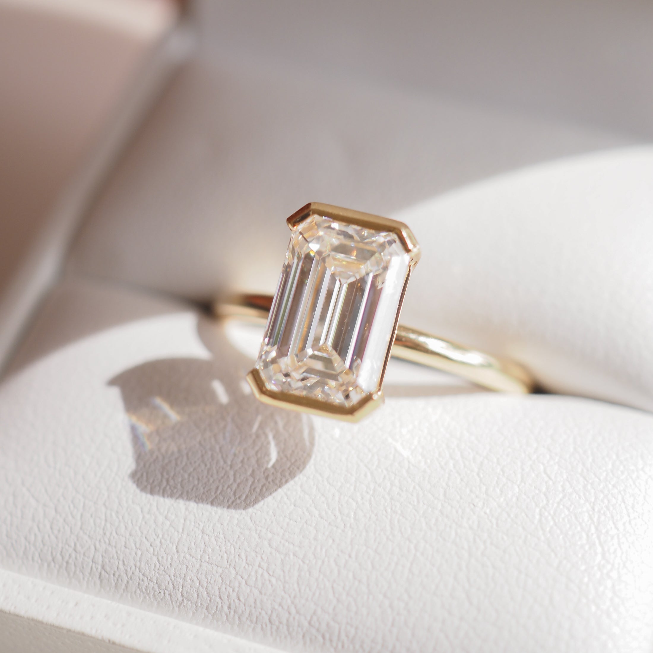 Emerald-cut diamond ring in gold setting, displayed on a white cushion with natural light highlighting its facets. Luxury jewellery.