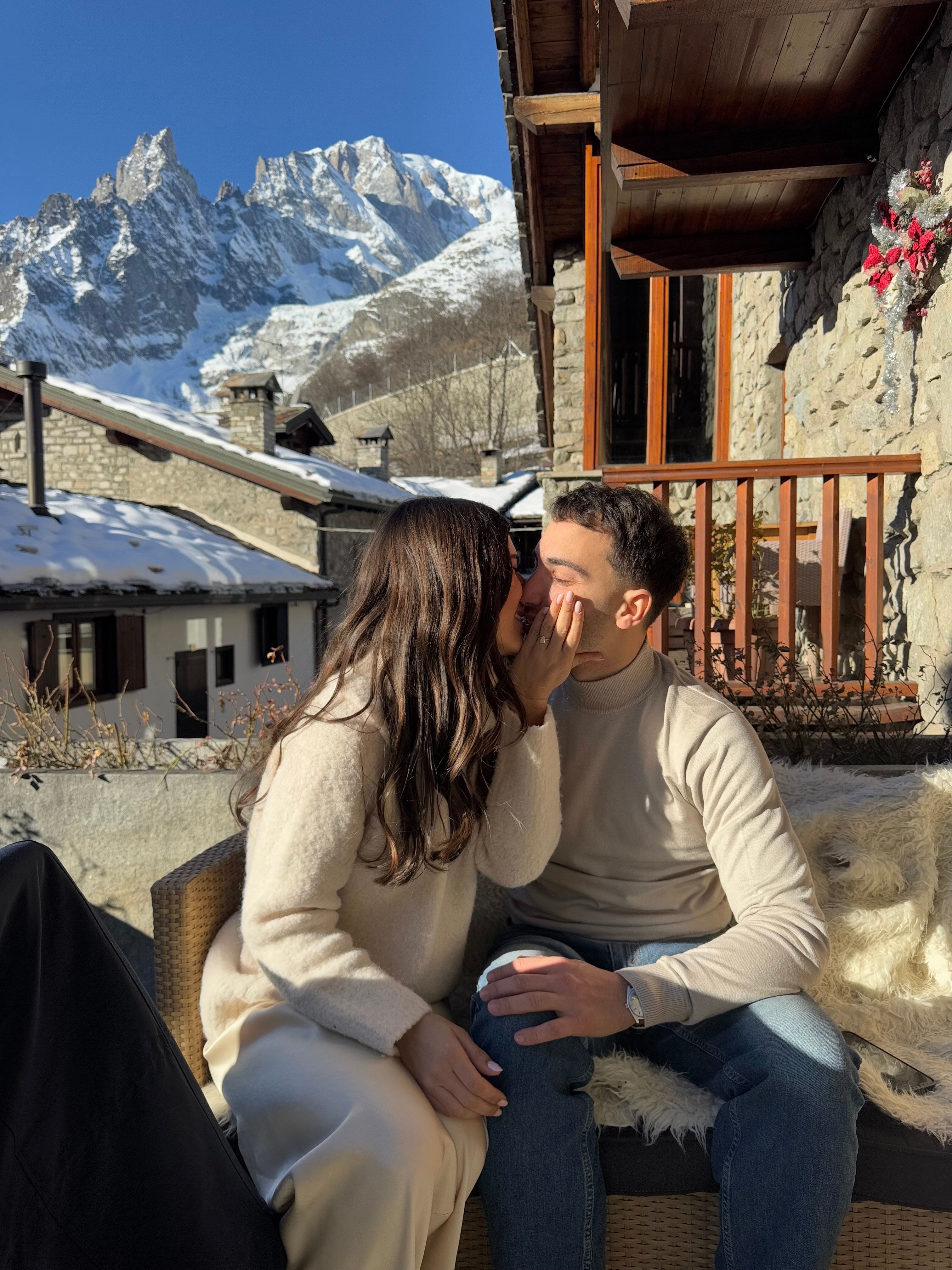 A couple sharing a secret on a snowy mountain terrace, surrounded by rustic wooden chalets, with a stunning backdrop of snow-capped peaks.