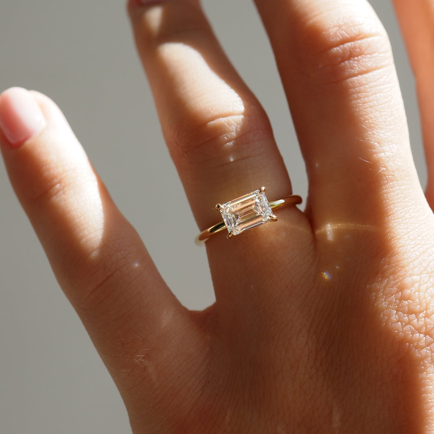 Hand wearing a gold band ring with a large emerald-cut diamond, reflecting sunlight. Luxury jewellery, elegant design.