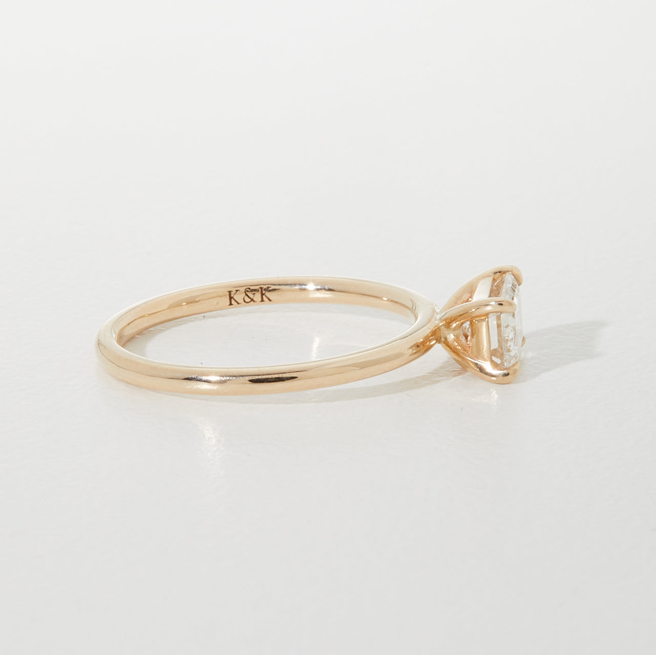 A rose gold engagement ring with a solitaire diamond in a minimalist setting. Engraved with "K&K" inside the band on a light background.