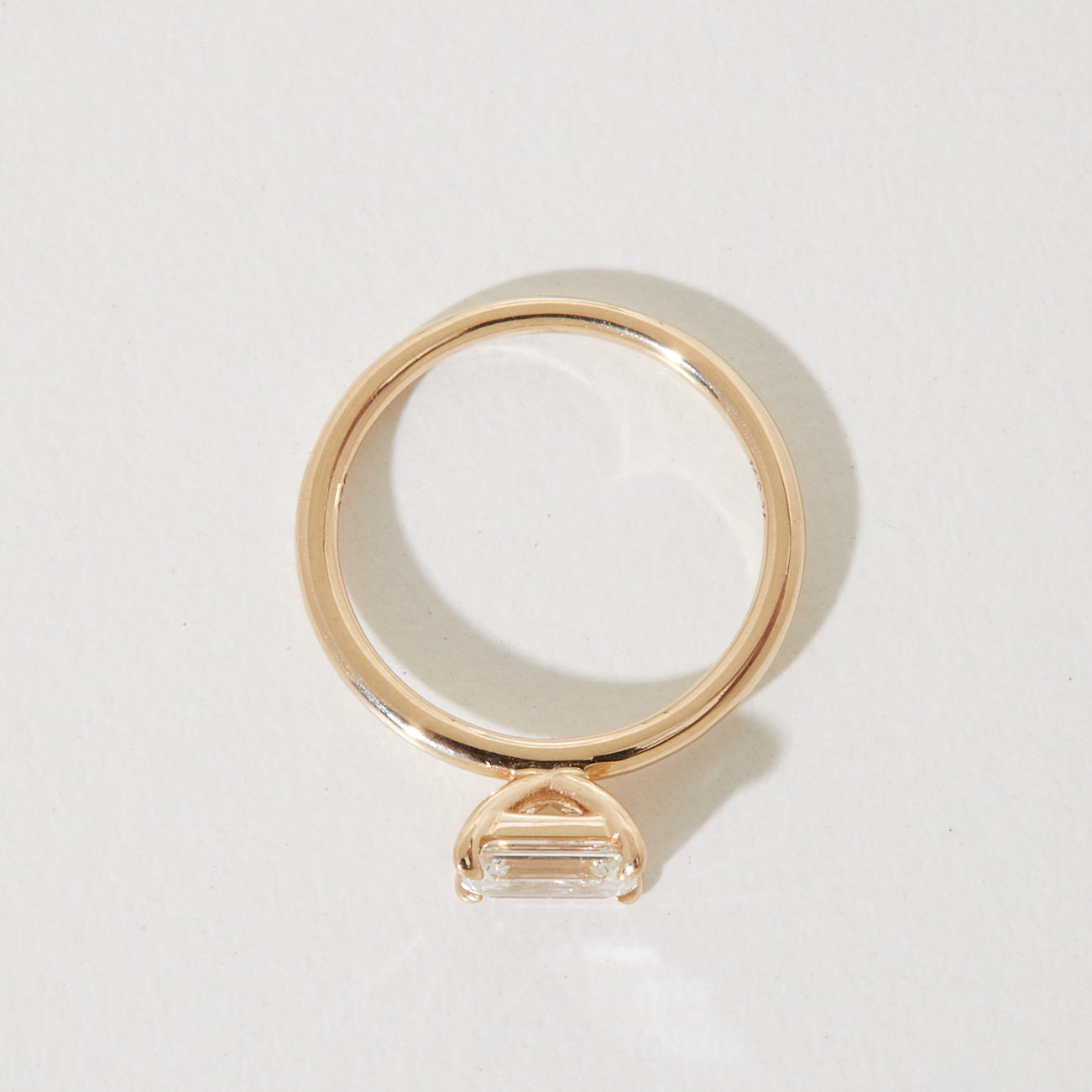 Gold ring with a horizontally-set emerald-cut diamond, casting a heart-shaped shadow on a light surface. Elegant luxury jewellery.
