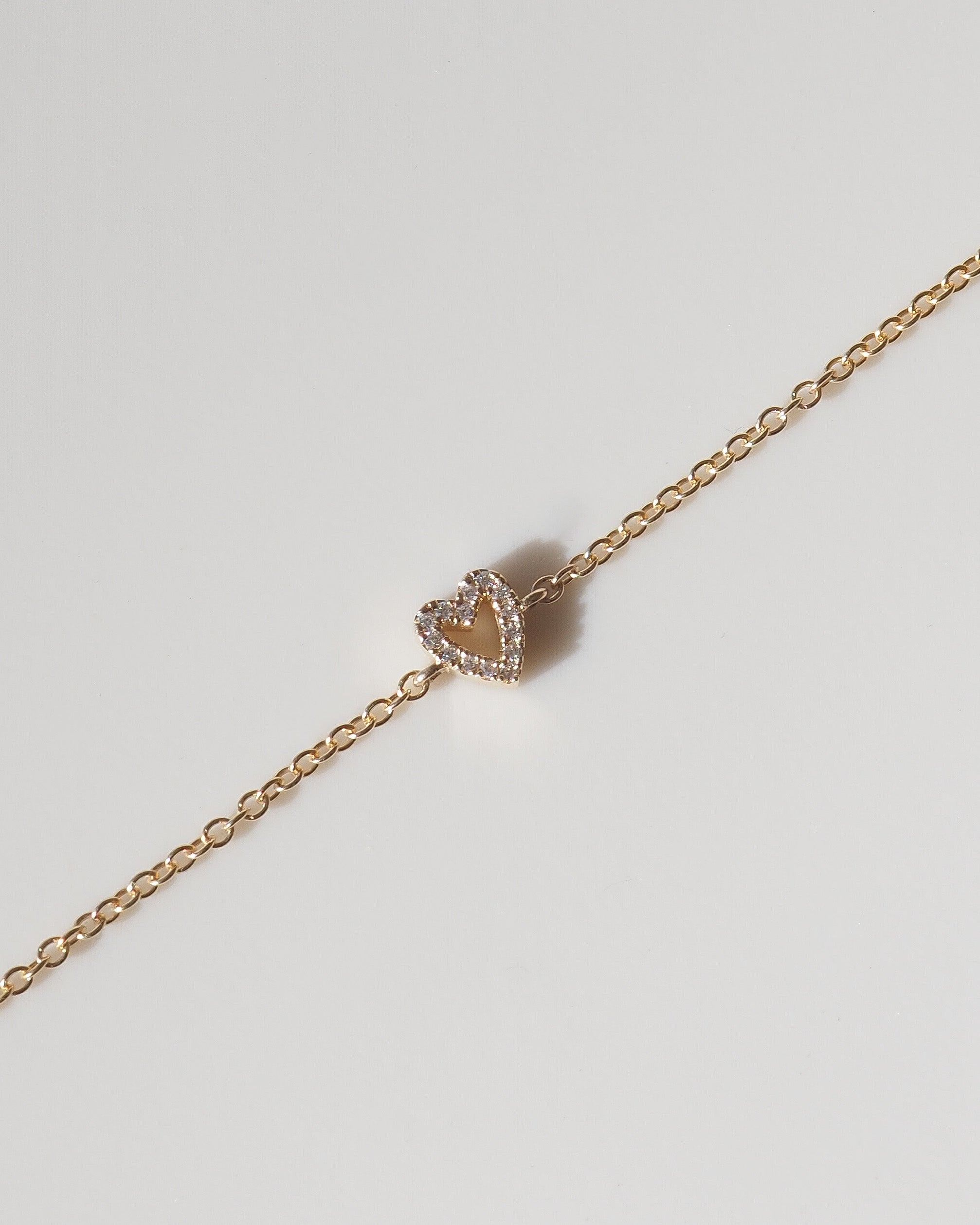 A delicate rose gold chain bracelet featuring a small heart-shaped charm with a double halo of clear stones, displayed diagonally on a light gray background.