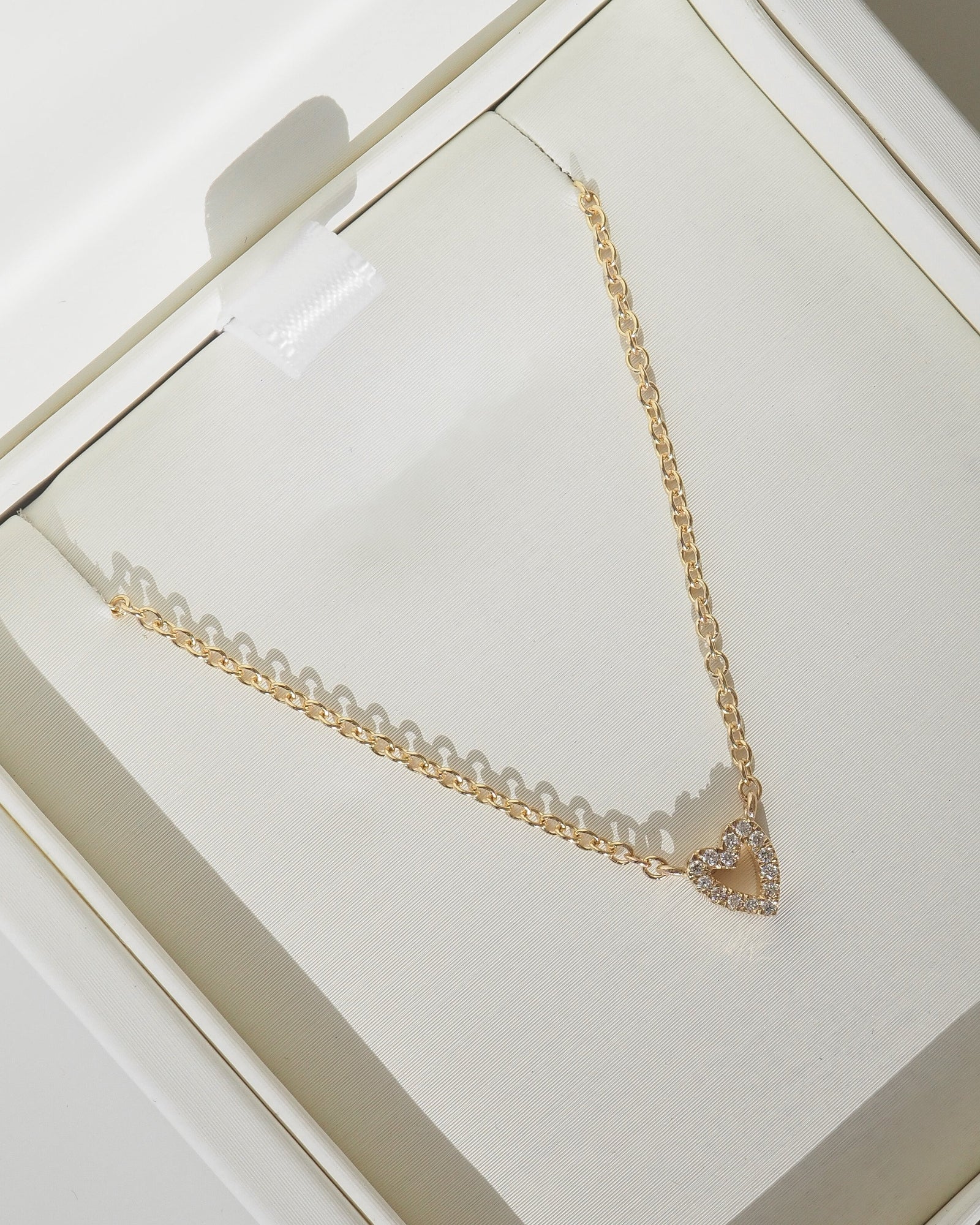 A gold chain necklace with a small heart-shaped pendant paved with clear stones, displayed inside a cream-colored jewelry box.