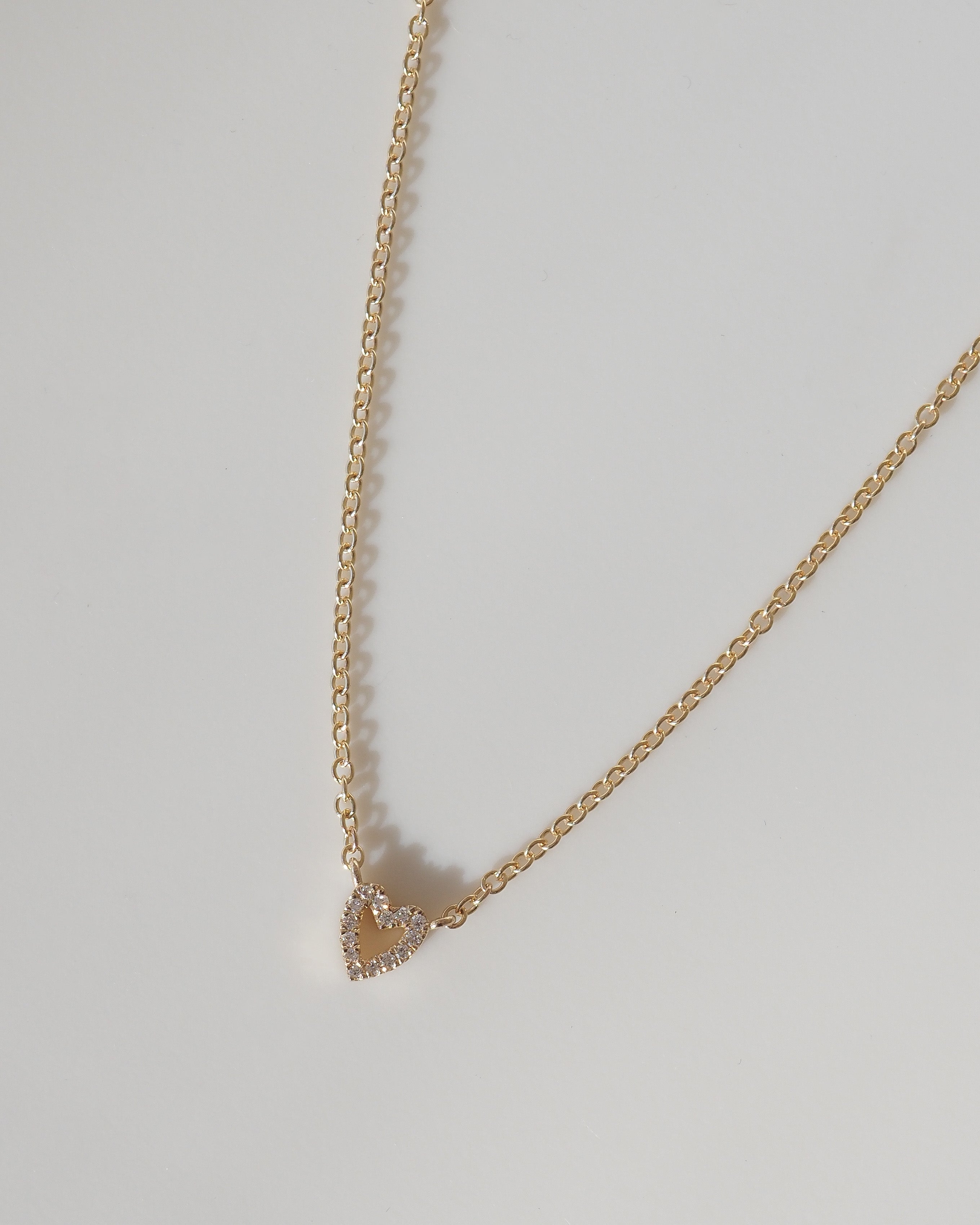 A delicate gold chain necklace with a small, openwork heart-shaped pendant studded with tiny clear crystals, displayed on a neutral light gray background.