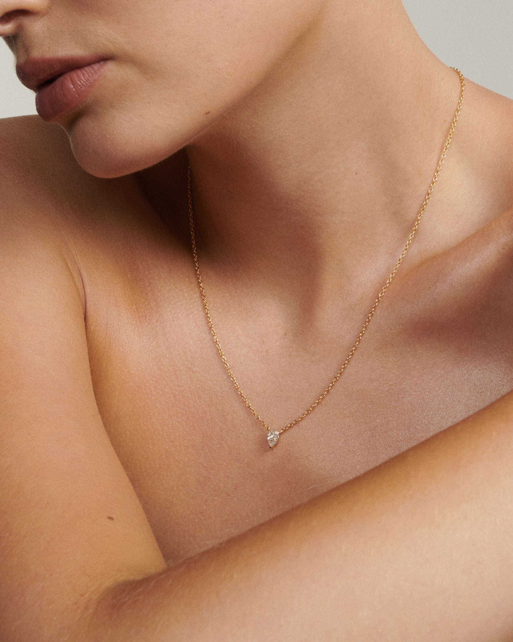 Gold necklace with a small diamond pendant on a person's neck [text overlay: Andrea wears the 0.5ct Pear Diamond Necklace]