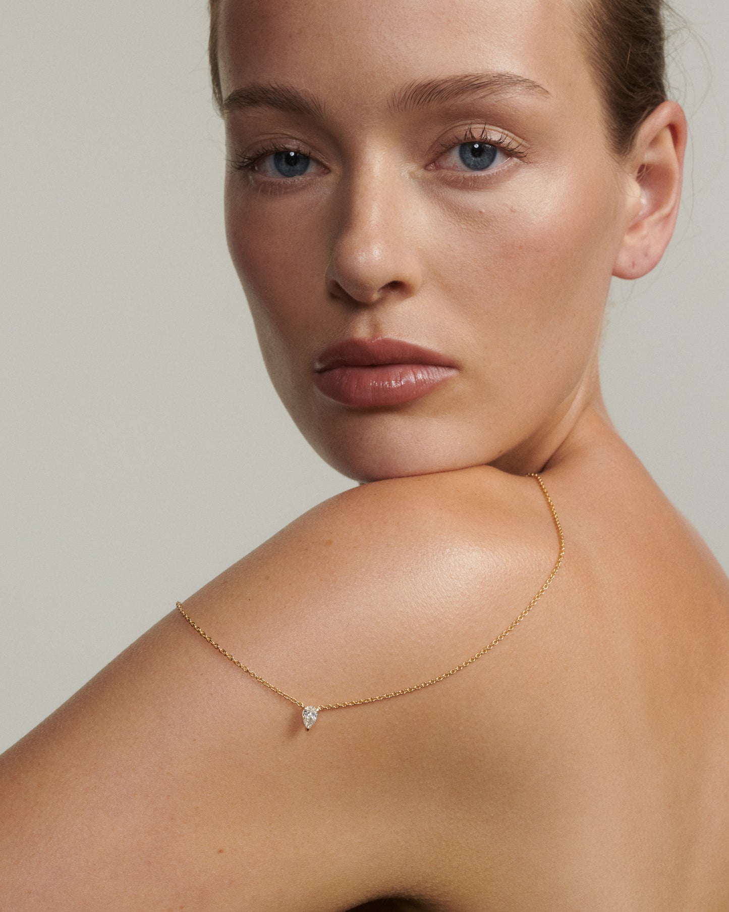 Woman wearing a delicate gold necklace with a small diamond pendant on a neutral background