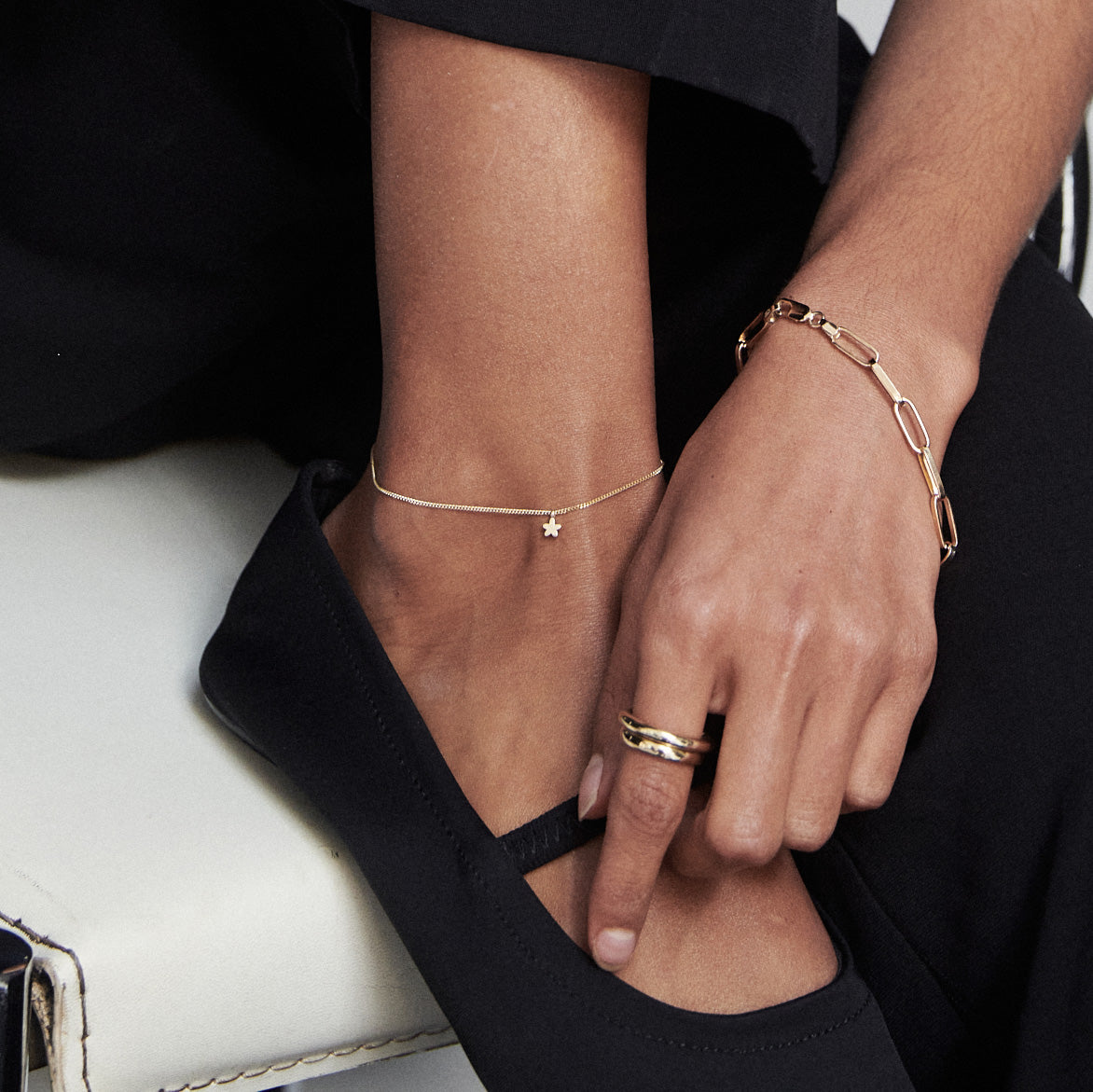 Close-up of an ankle and hand adorned with gold jewellery: a delicate anklet with a star charm, a chain bracelet, and stacked rings. Black shoes visible.