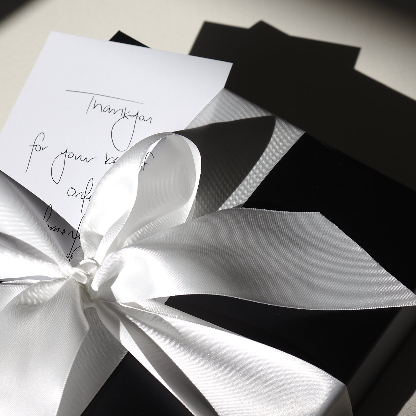 Gift box with elegant white satin ribbon and a handwritten thank-you note, suggesting luxury jewellery packaging in soft natural light.