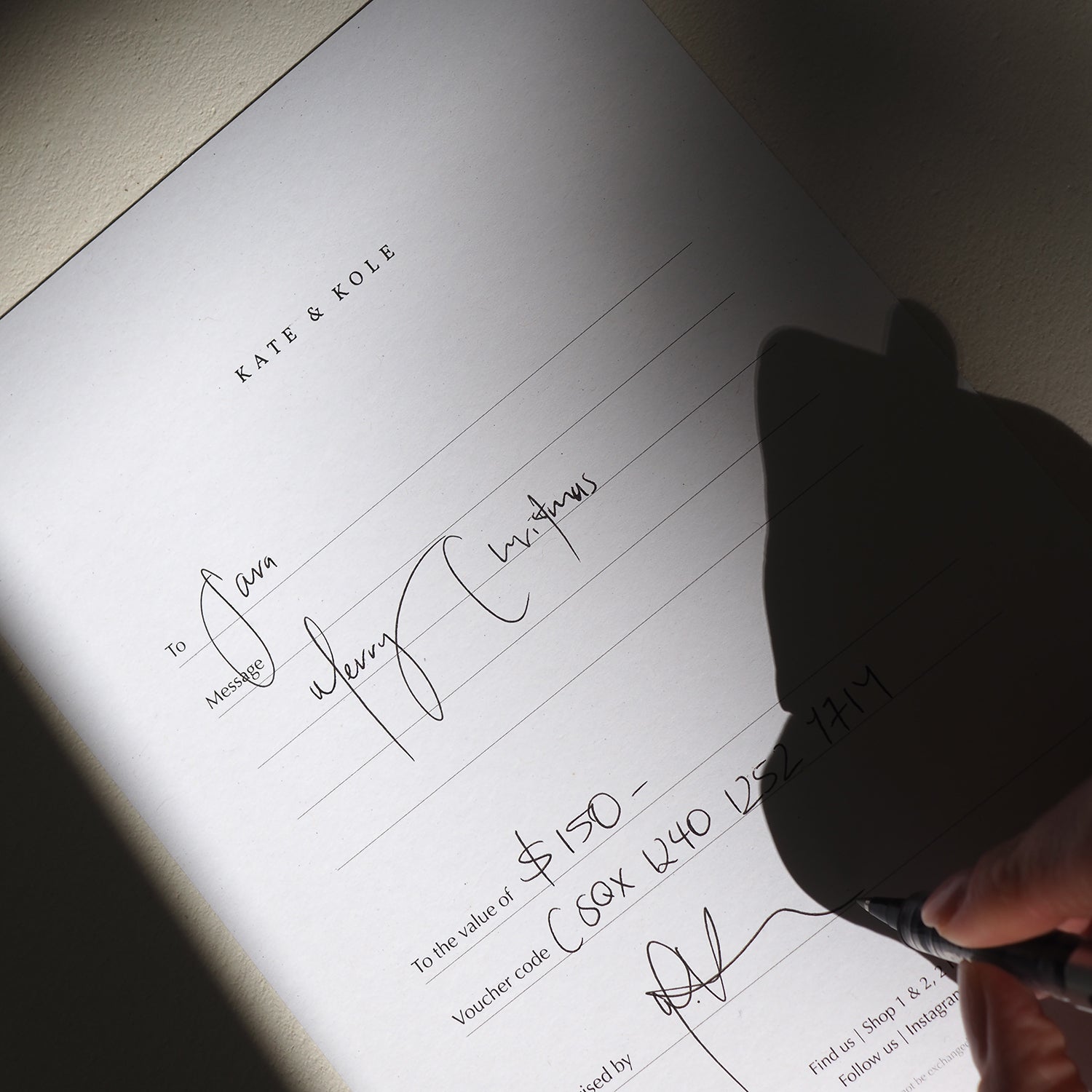 Hand signing a luxury jewellery gift voucher from Katie & Kole, reading "Merry Christmas" valued at $150, on a well-lit surface.