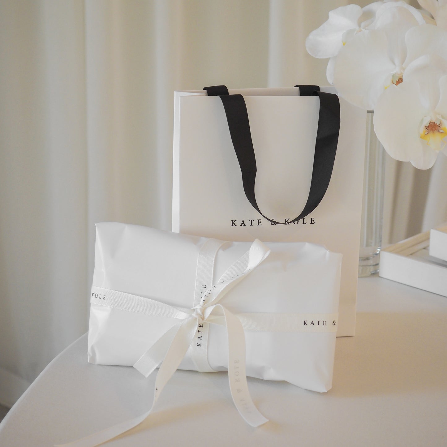 Luxury jewellery gift set with elegant white packaging and black ribbon by Kate & Kole, placed on a table with orchids.