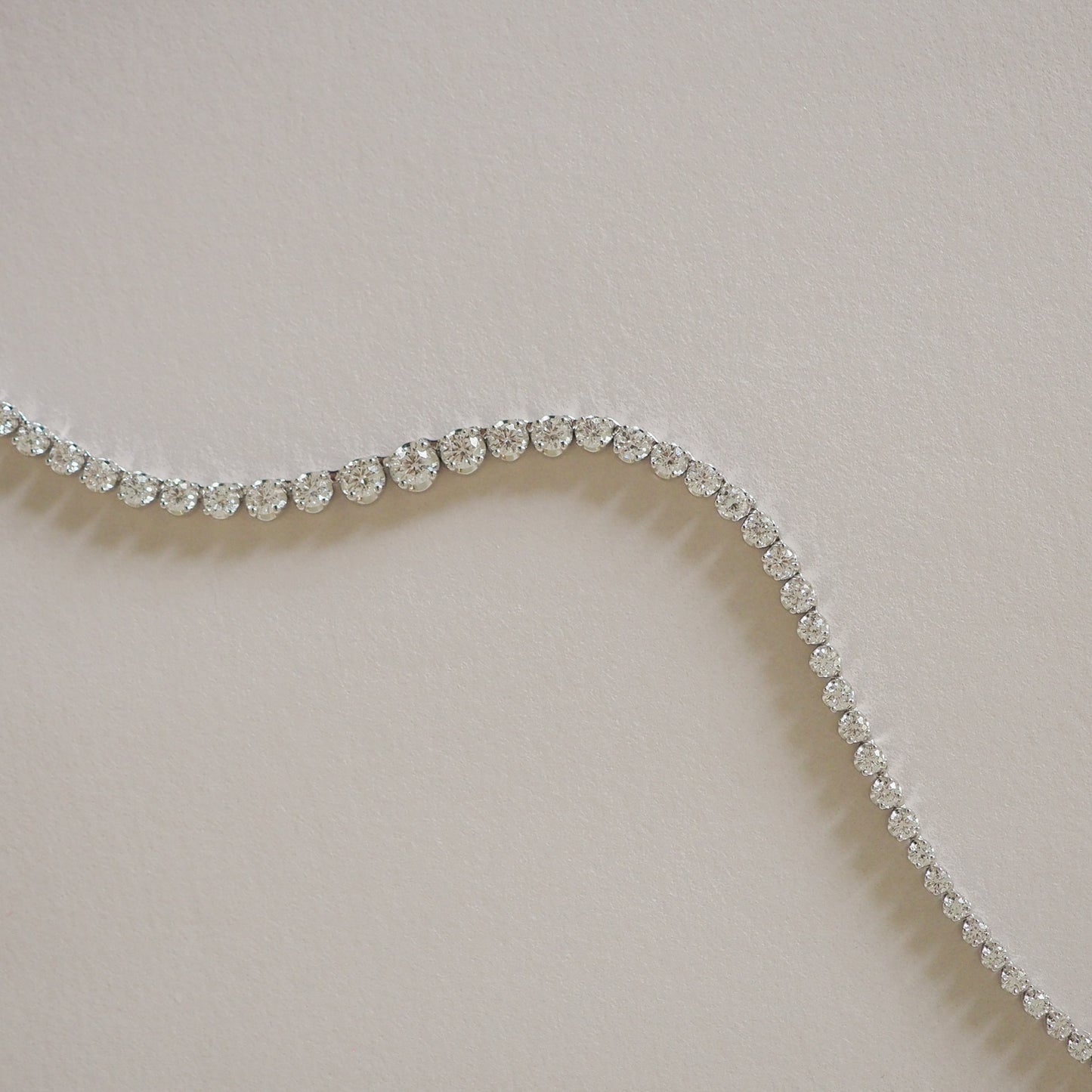 Elegant diamond tennis bracelet on a subtle beige background, showcasing luxury jewellery with a flowing, curved design.