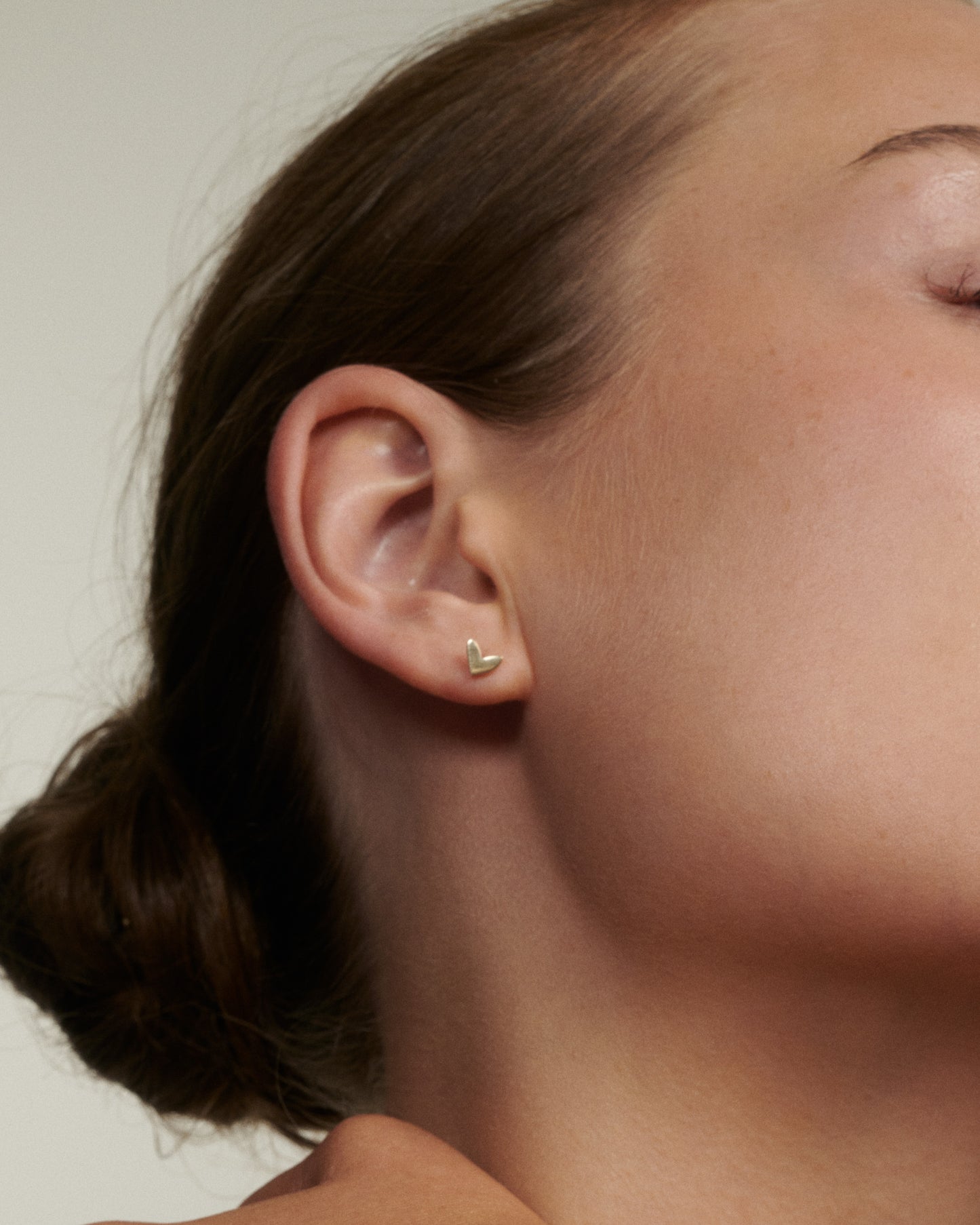 Close-up of a person wearing a gold heart-shaped earring against a neutral background