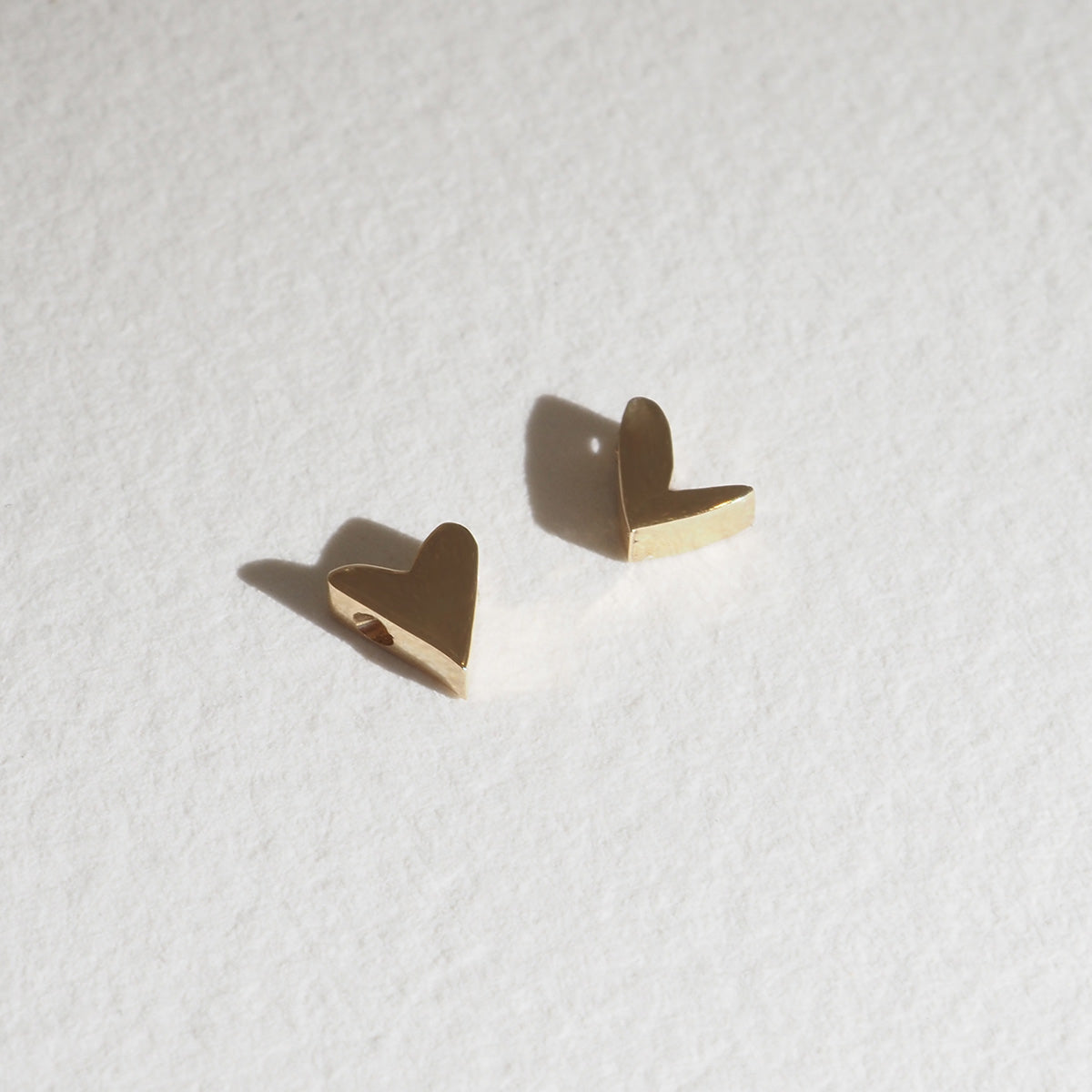 Gold heart-shaped stud earrings on textured white background, casting soft shadows. Luxury jewellery.