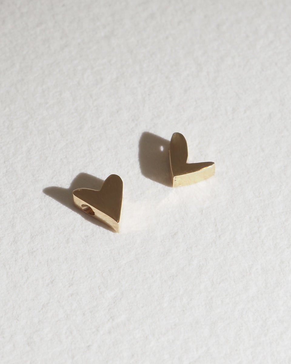 Gold heart-shaped stud earrings on textured white background, casting soft shadows. Luxury jewellery.