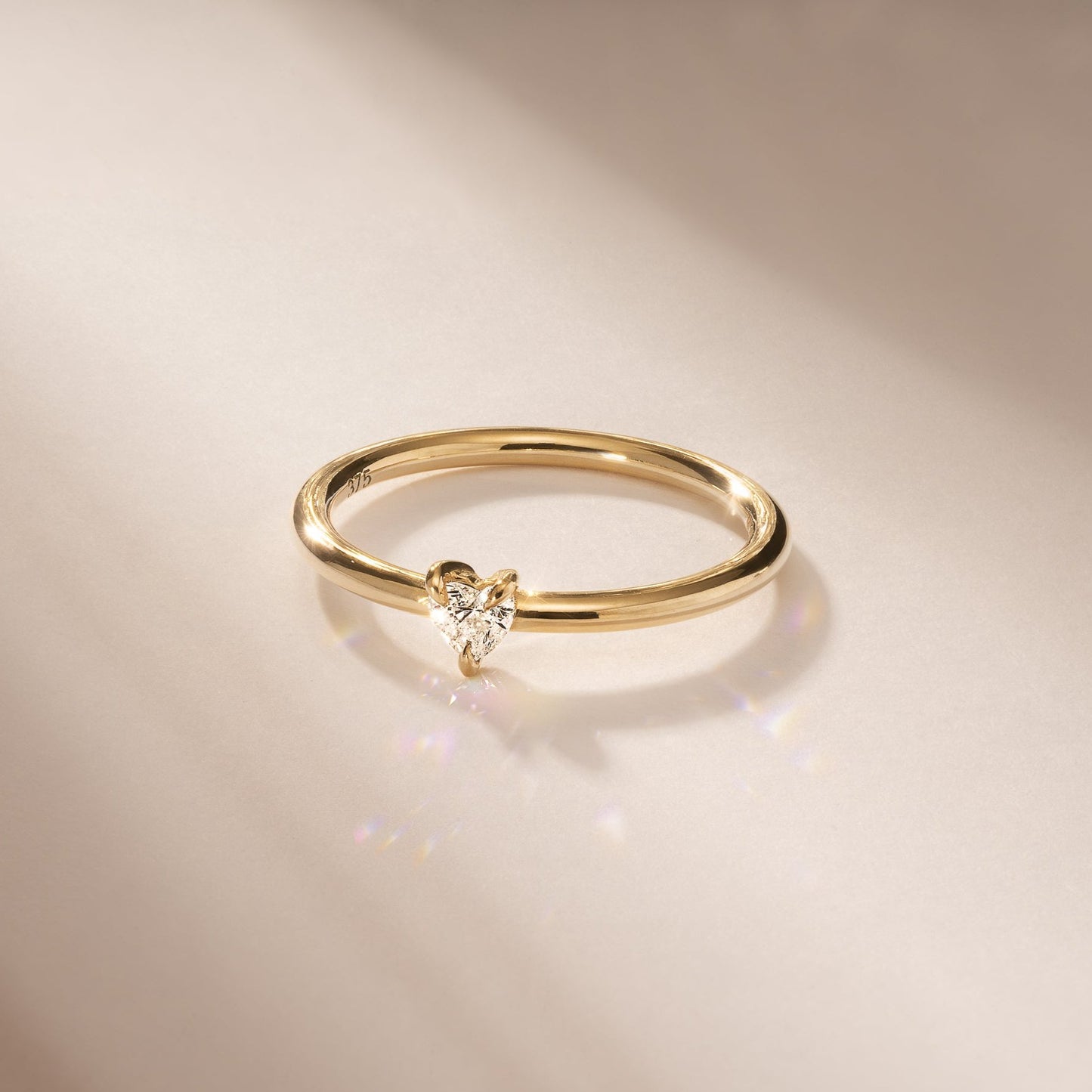 Gold ring with a heart-shaped diamond on a smooth, beige surface; elegant luxury jewellery piece reflecting light subtly.