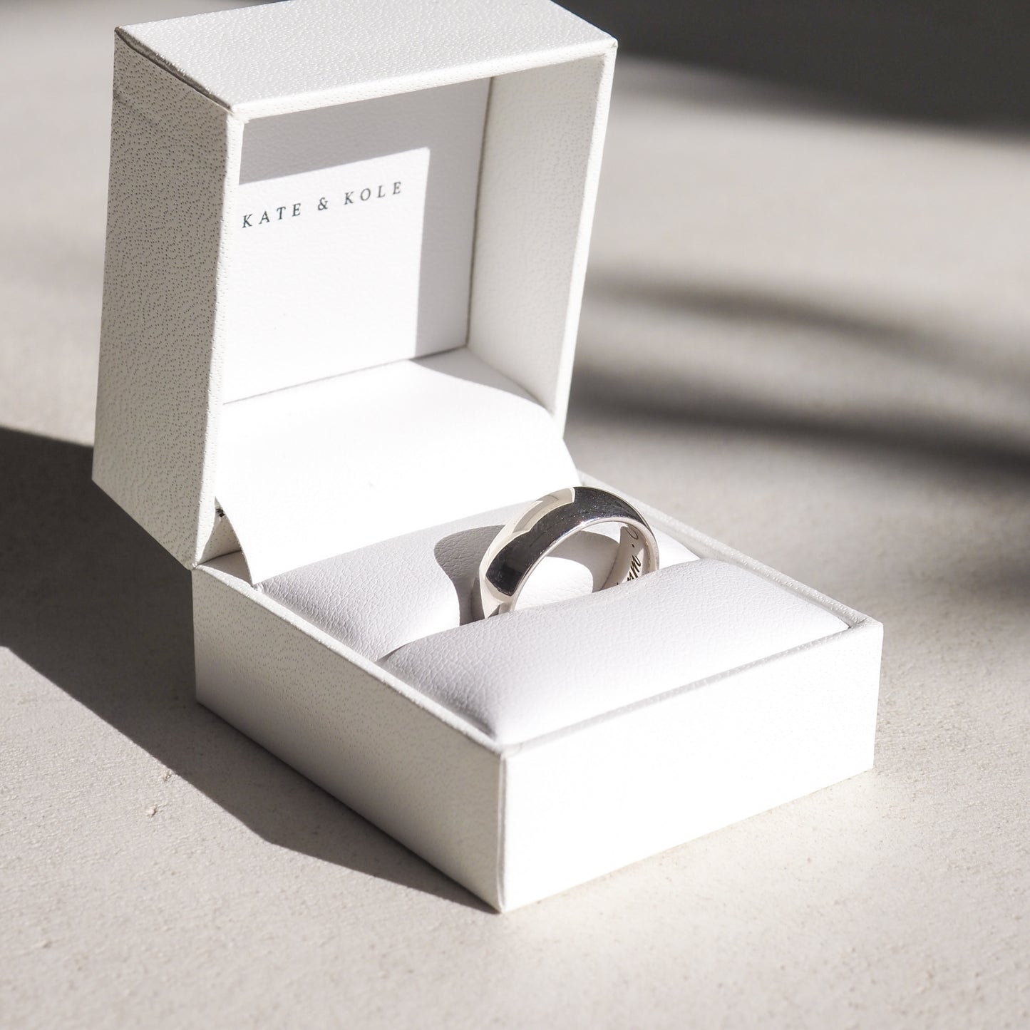 Silver ring elegantly displayed in a white Kate & Kole jewellery box, casting soft shadows on a light surface.