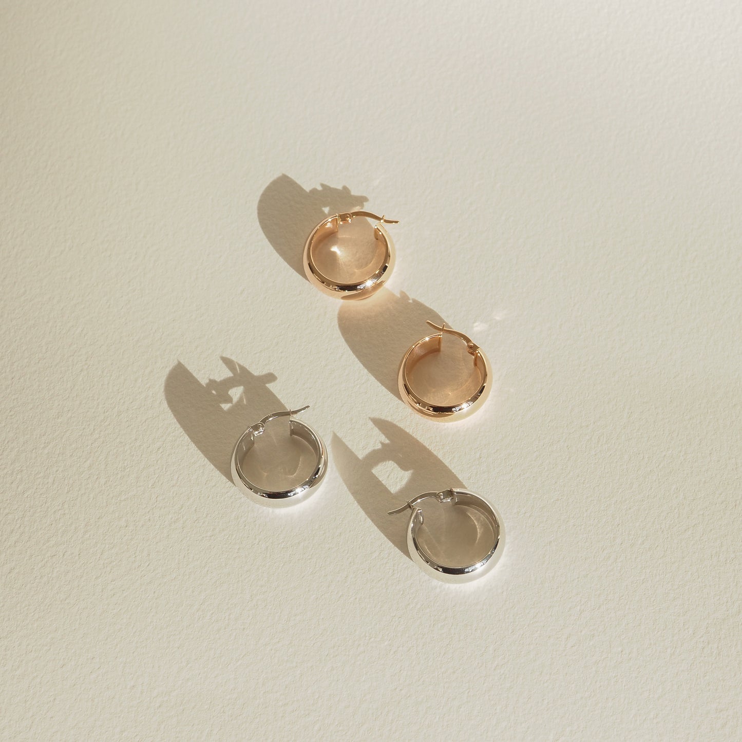 Two pairs of hoop earrings, one gold and one silver, lie on a light surface casting subtle shadows. Elegant, minimalist jewellery display.