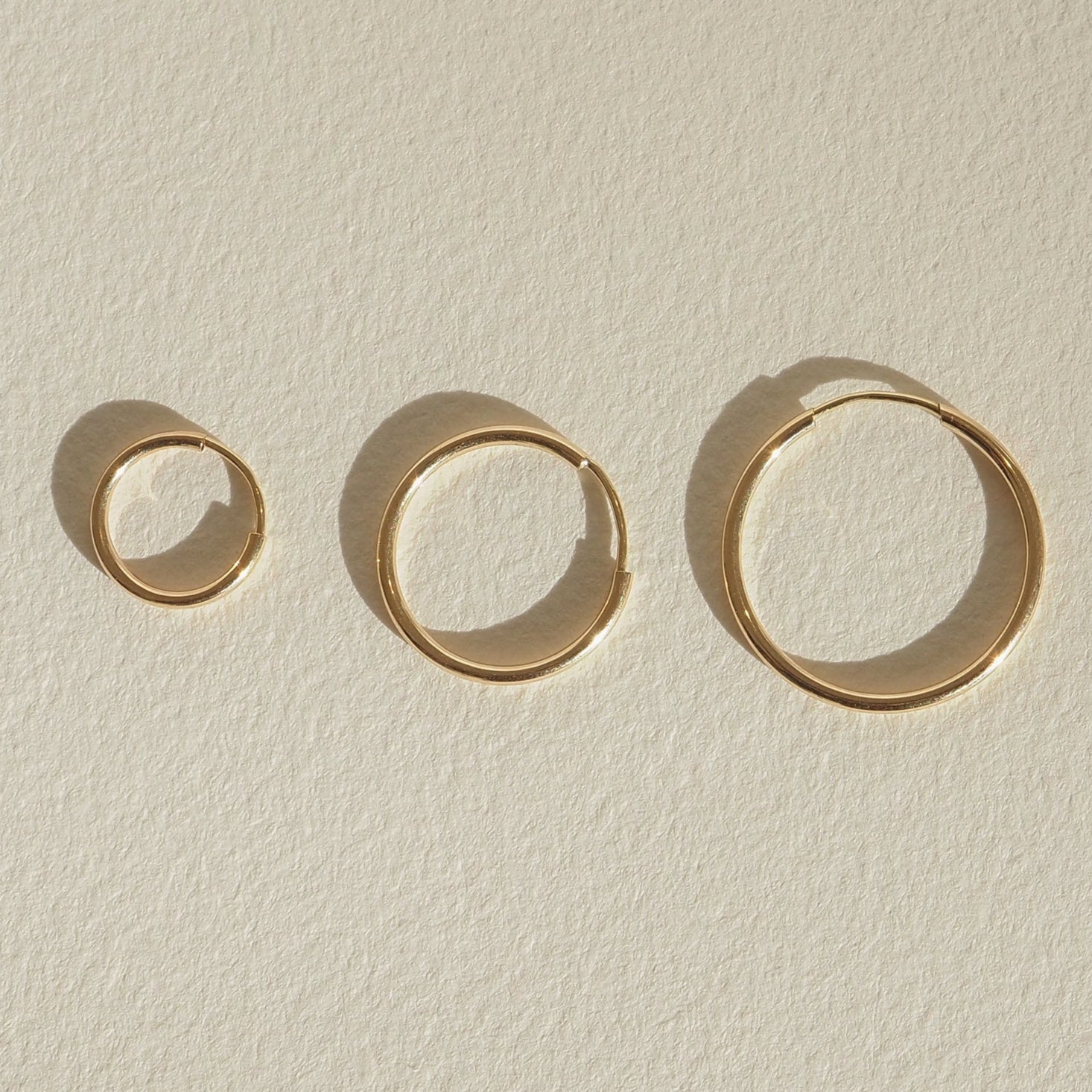 Four gold bangles of varying sizes displayed on a light textured surface, casting soft shadows, showcasing minimalist luxury jewellery design.