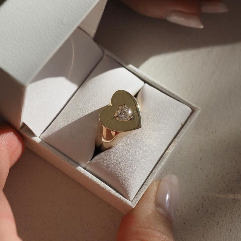 Gold ring with heart-shaped face, featuring a central diamond, presented in a white jewellery box. Perfect for luxury engagements.