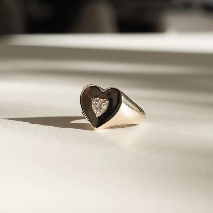 Gold signet ring with black heart emblem, featuring a small diamond in the centre, resting on a softly lit surface.