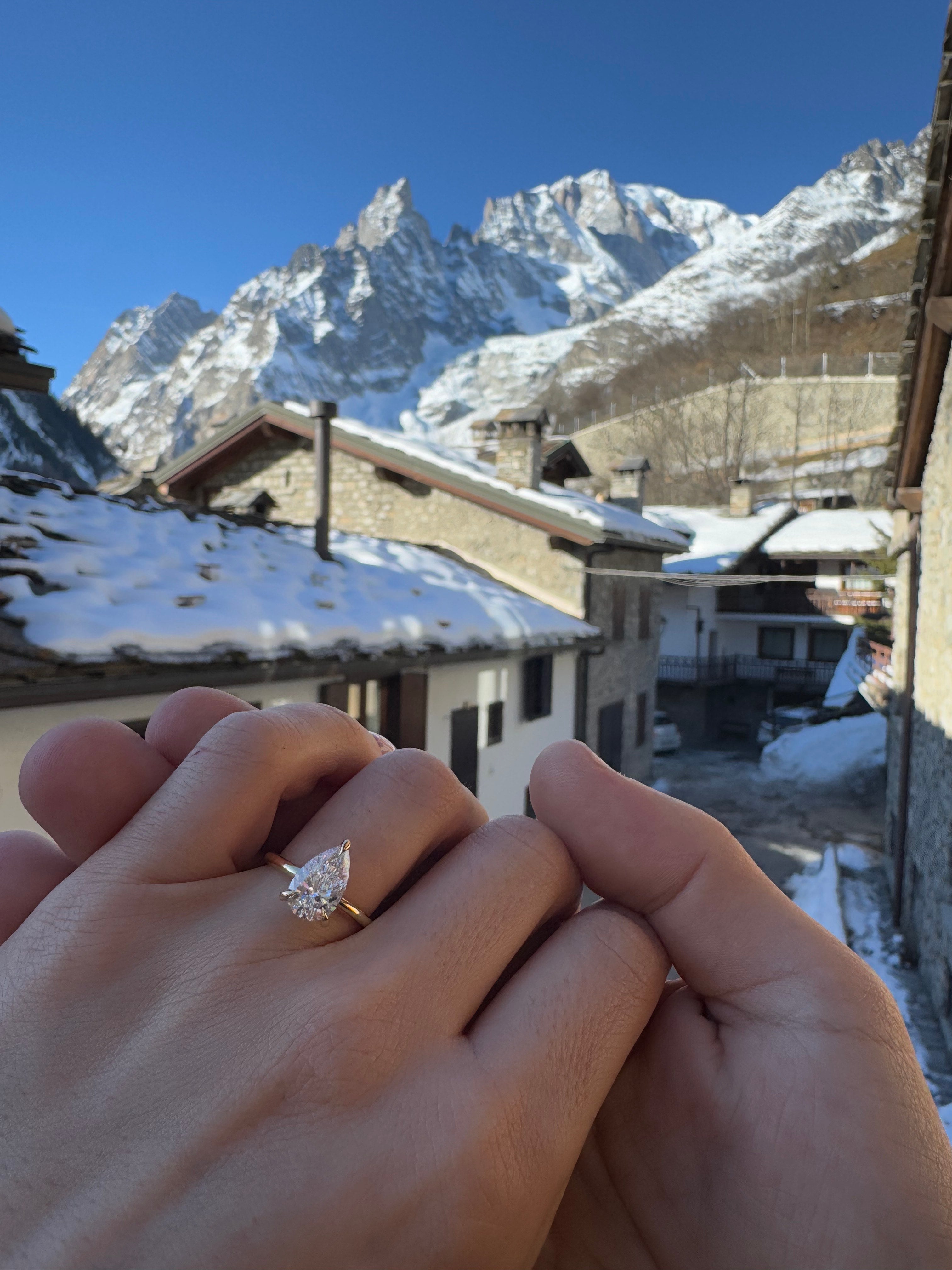 A hand wearing a pear-cut diamond ring with mountain village backdrop, snow-covered roofs, and clear blue sky. Luxury jewellery, alpine scenery.