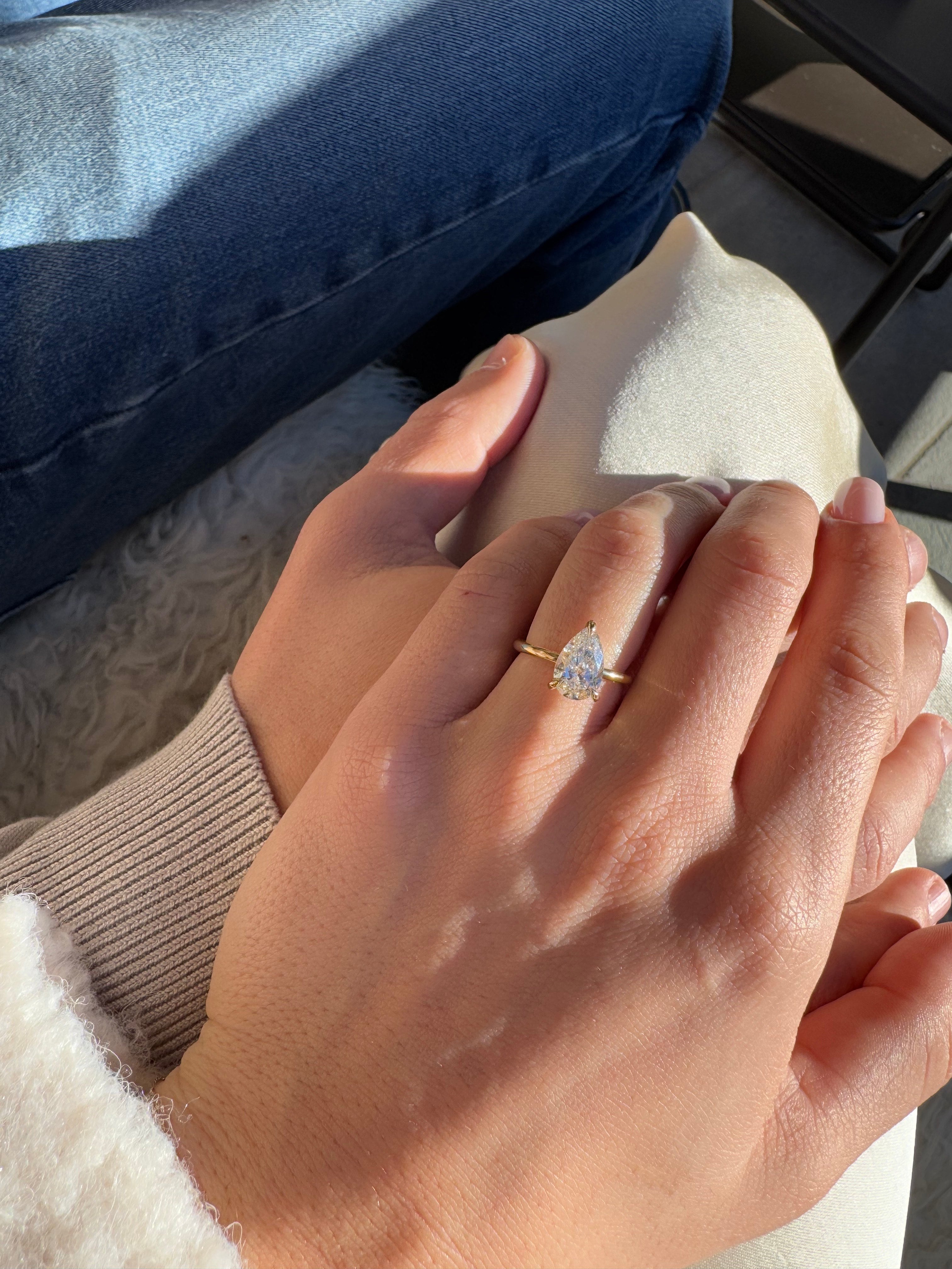 Close-up of hands resting on a lap, highlighting a pear-shaped diamond engagement ring on a gold band, with soft sunlight casting shadows.