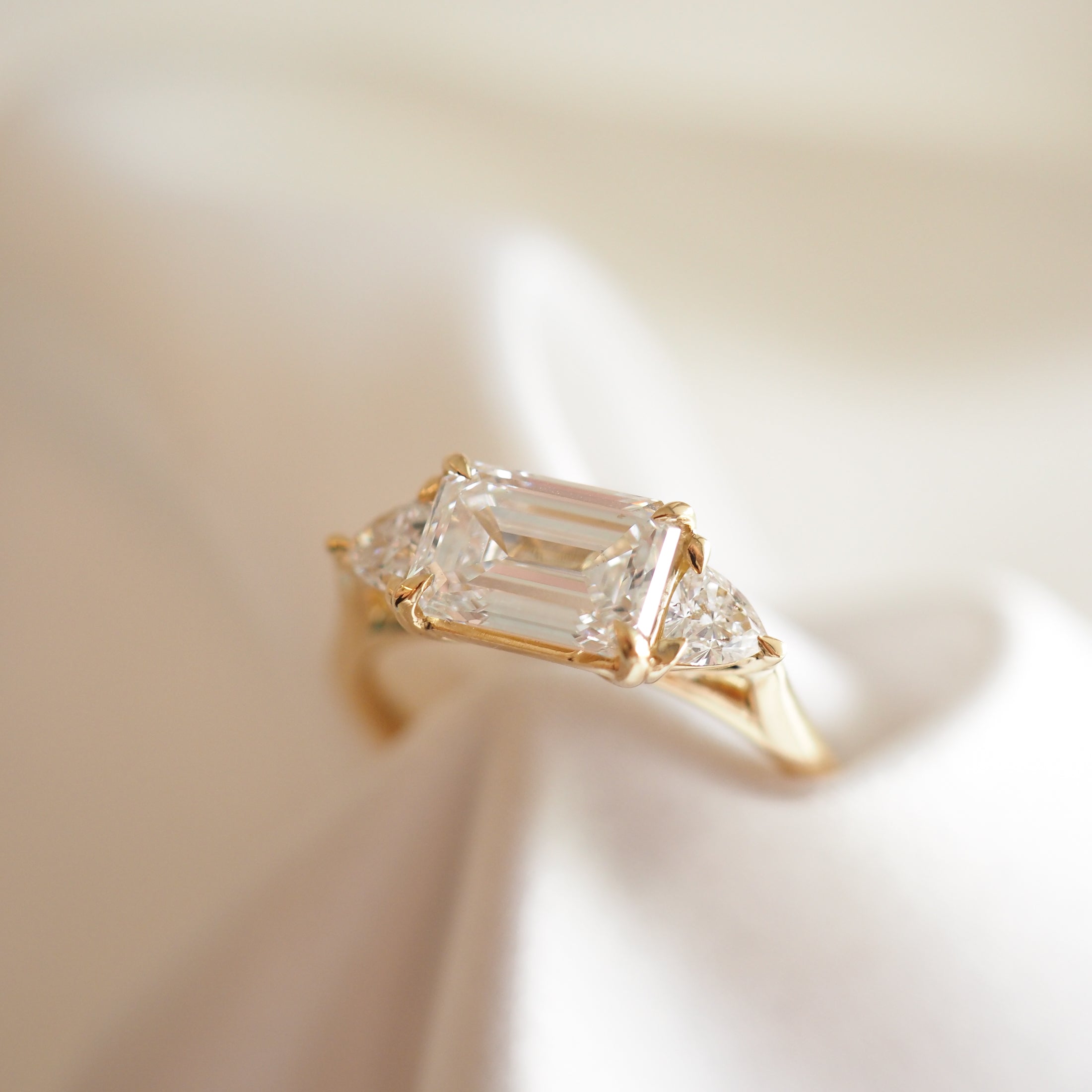Emerald-cut diamond ring with two side stones on a gold band, elegantly displayed on a soft, white fabric background.