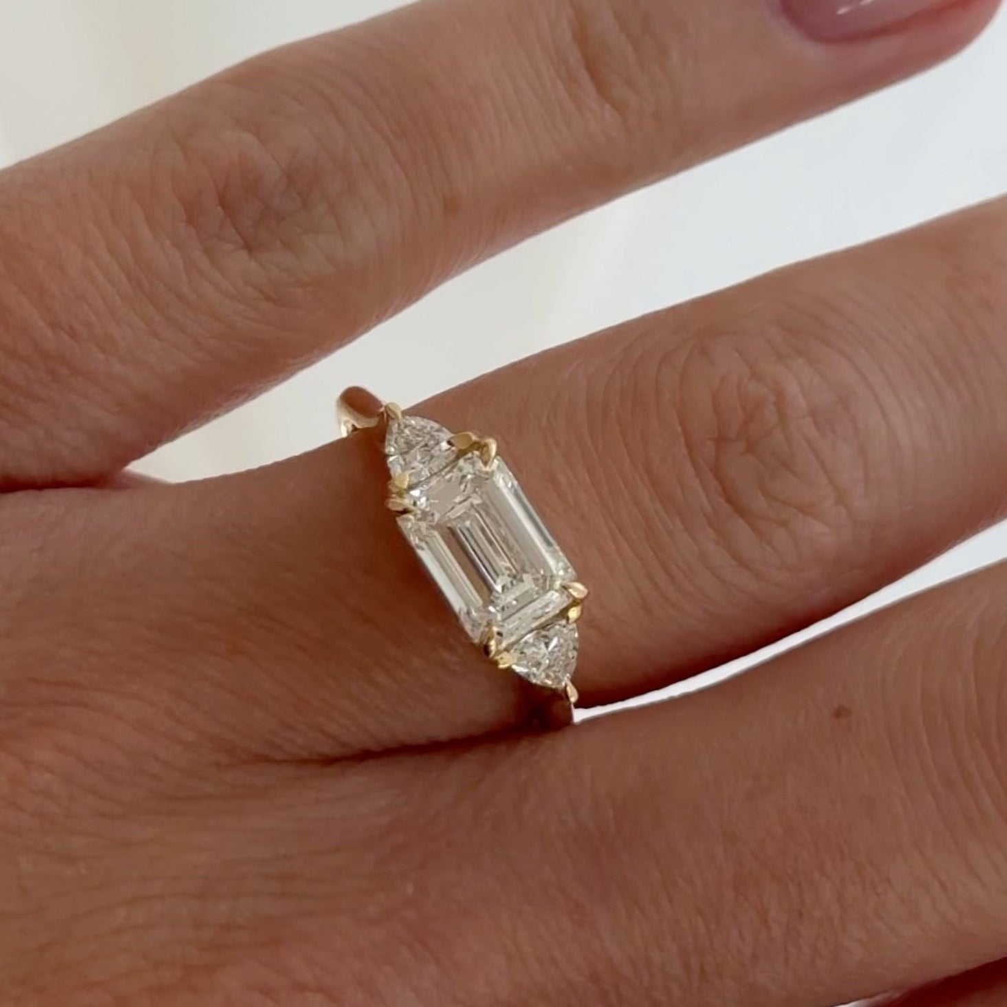 Close-up of a hand wearing a luxury engagement ring featuring an emerald-cut centre diamond flanked by two trapezoid diamonds on a gold band.