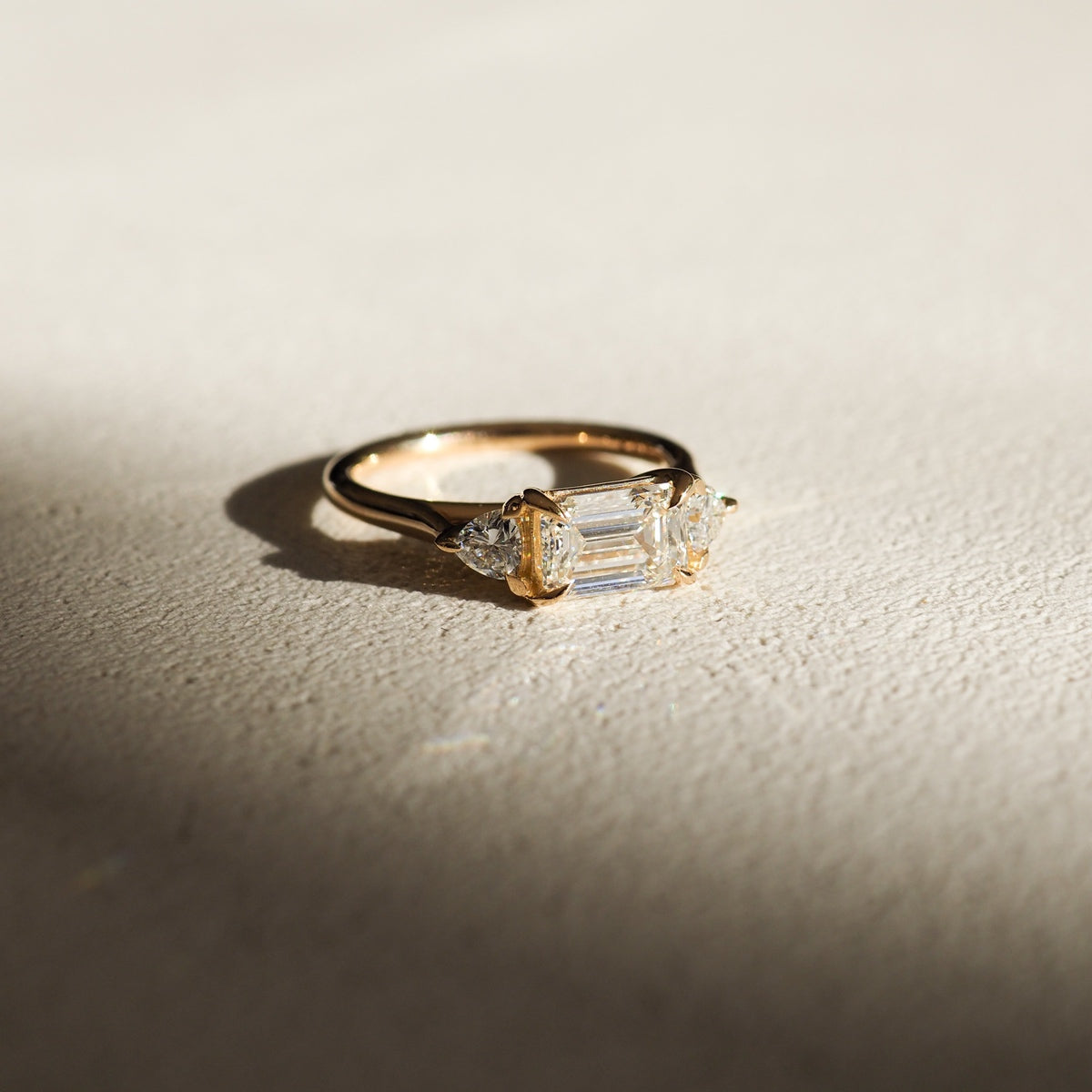 Gold ring with an emerald-cut diamond centre and two trilliant-cut diamonds on either side, casting a delicate shadow on a textured surface.