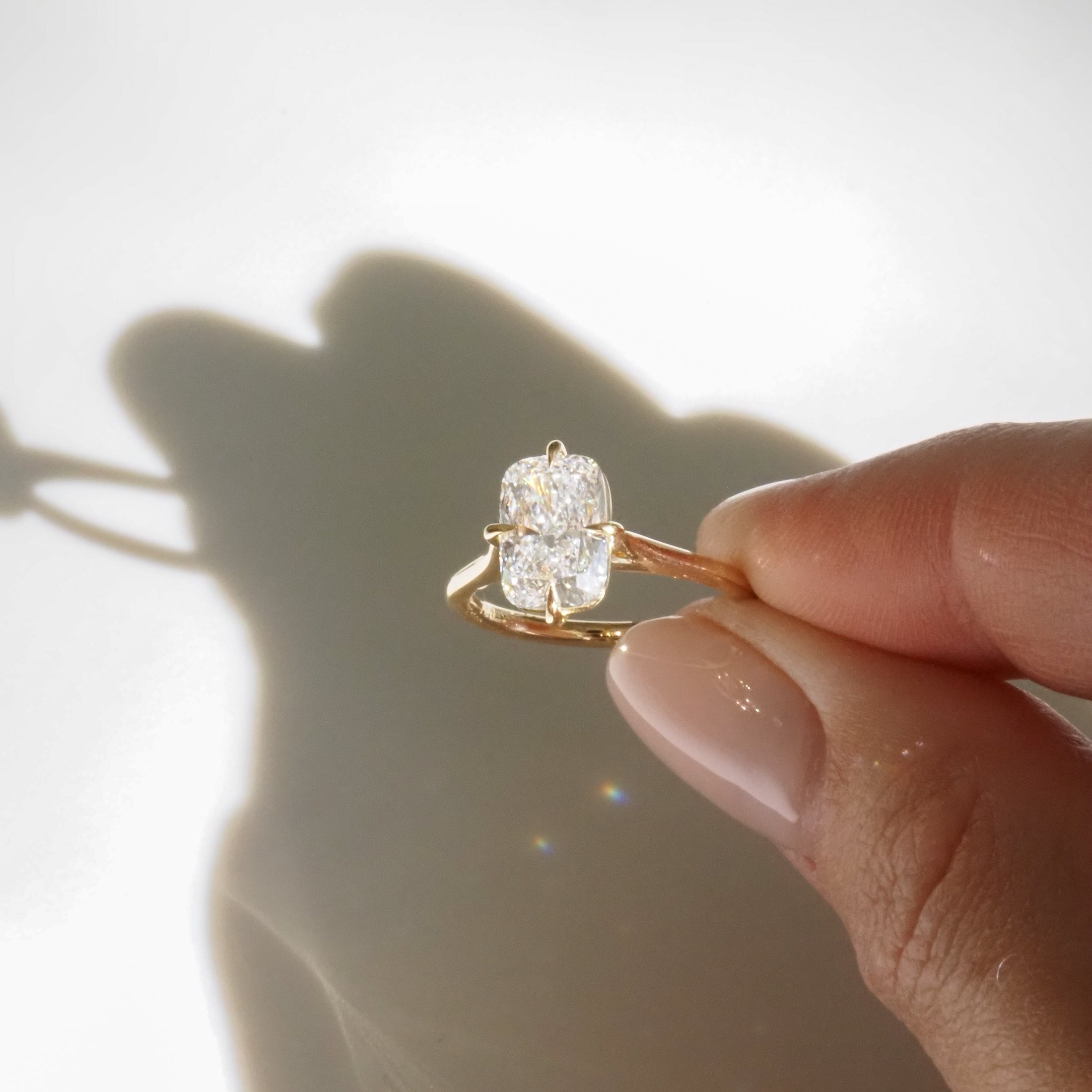 Hand holding a luxury gold ring with a large oval diamond, casting a shadow on a white surface, showcasing fine jewellery elegance.