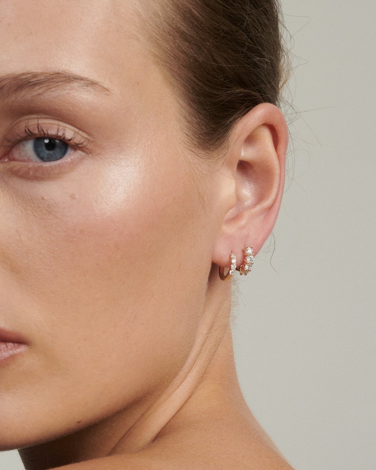 Close-up of a woman wearing earrings with a neutral background [text overlay: Tiny and Signature Diamond Huggies]