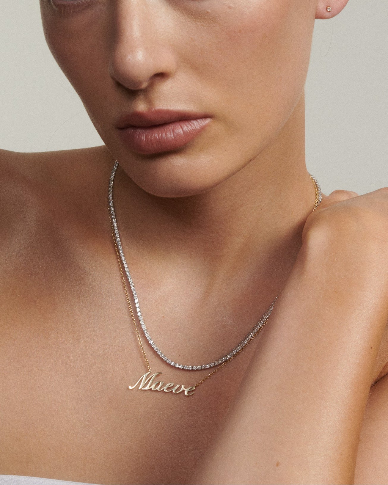 Woman wearing a gold necklace with 'Maeve' on a neutral background [text overlay: Andrea wears our Name Plate Necklace & Tennis Necklace]