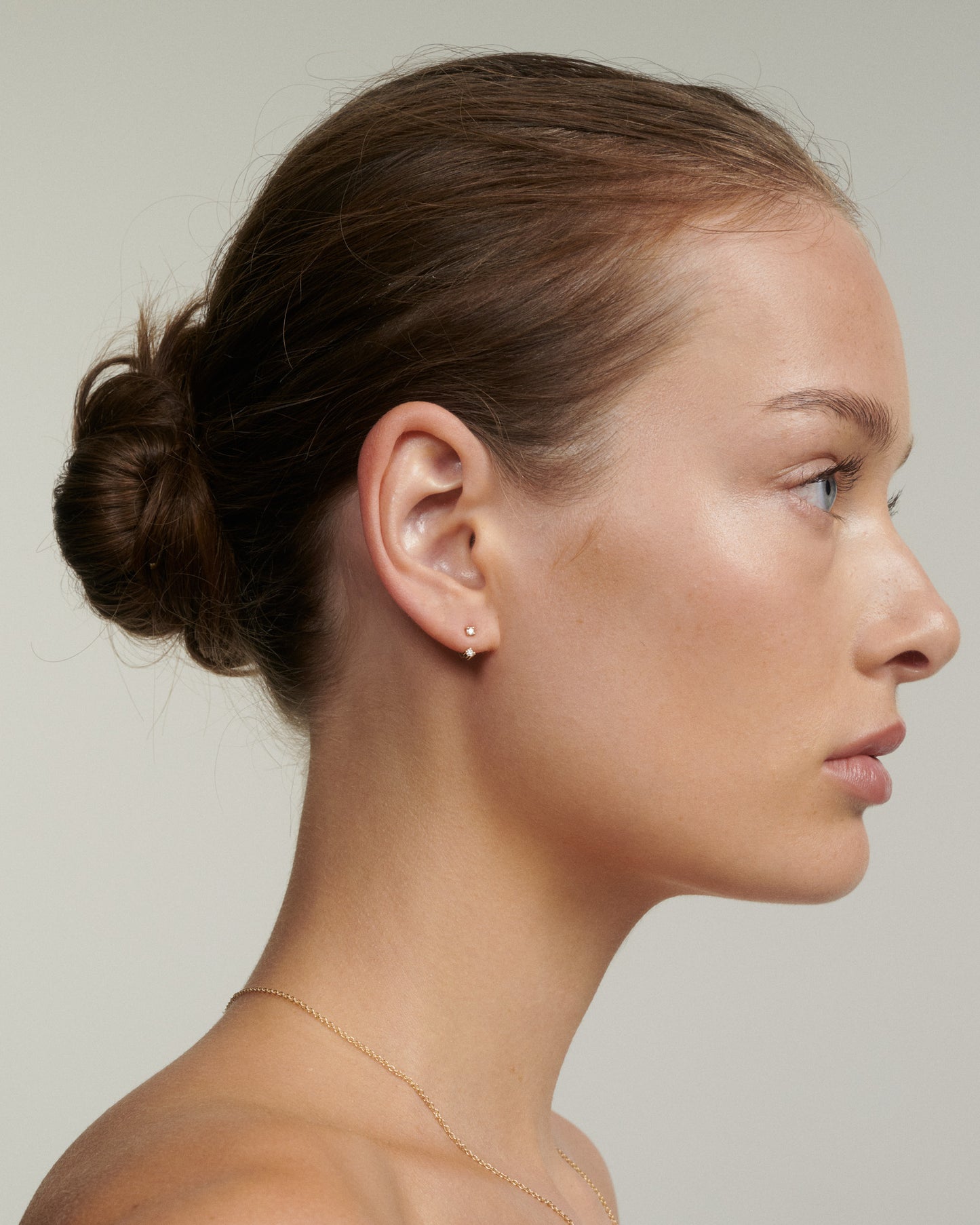 Side profile of a woman with a simple hairstyle and minimal jewelry against a neutral background