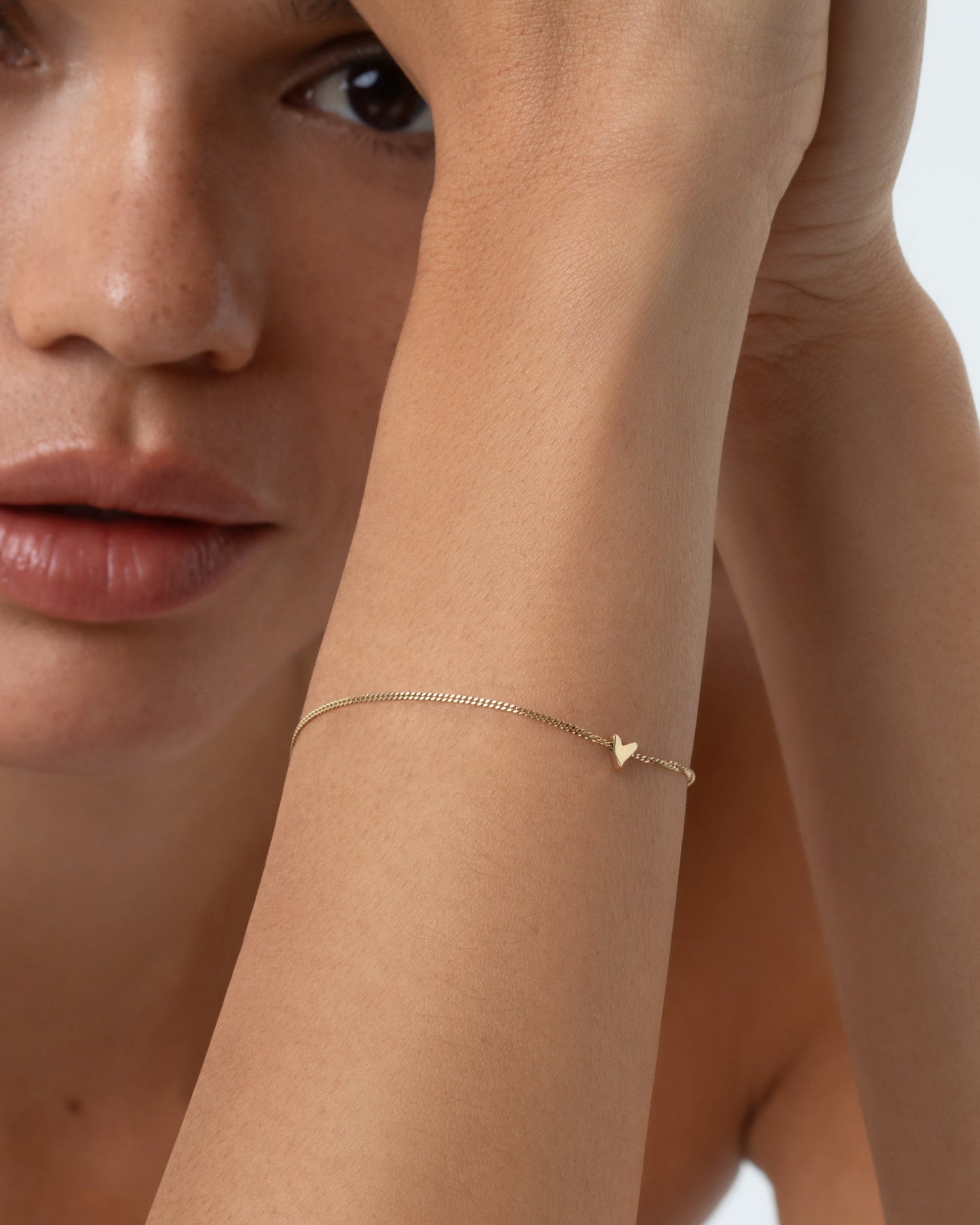 A close-up lifestyle shot of a model wearing a delicate gold chain bracelet and a thin gold bracelet with a small heart-shaped charm, posed against a clean white background.