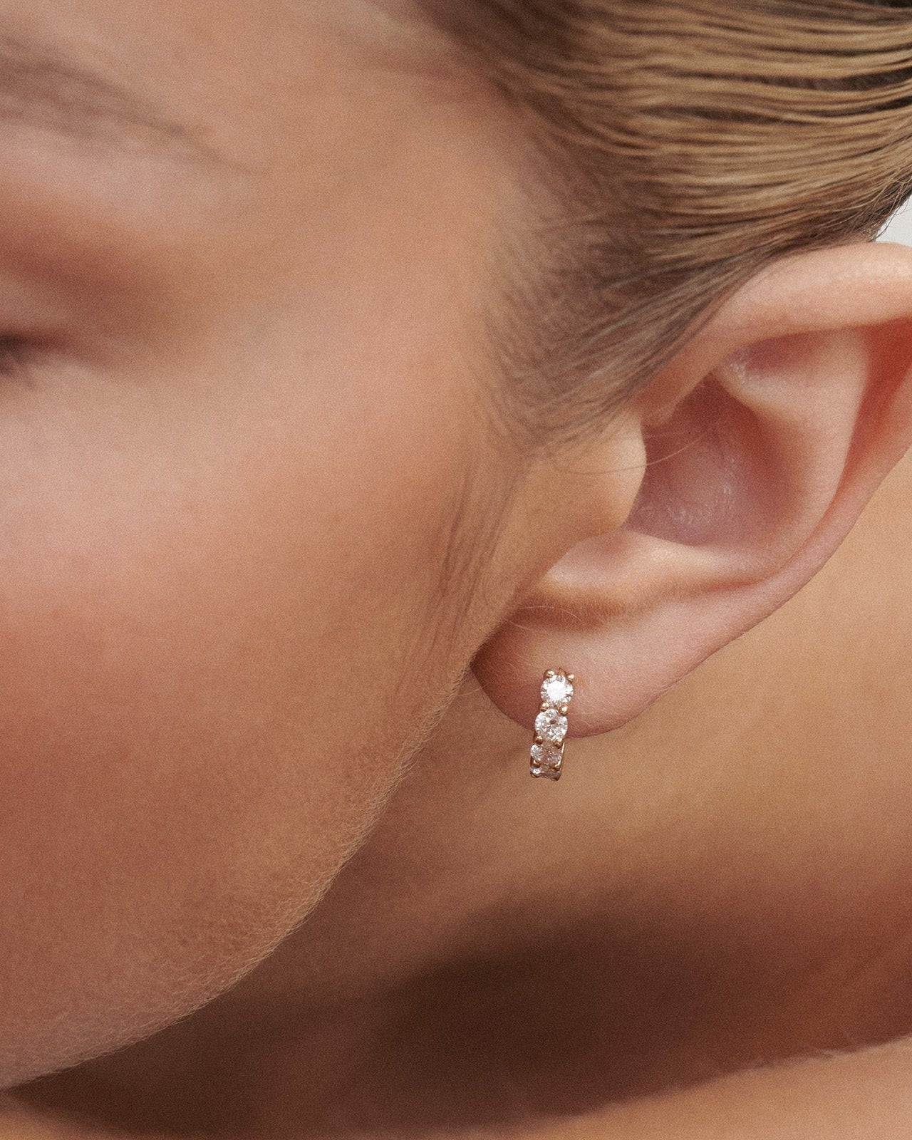 Close-up of a woman wearing a diamond earring. [text overlay: Signature Diamond Huggies]