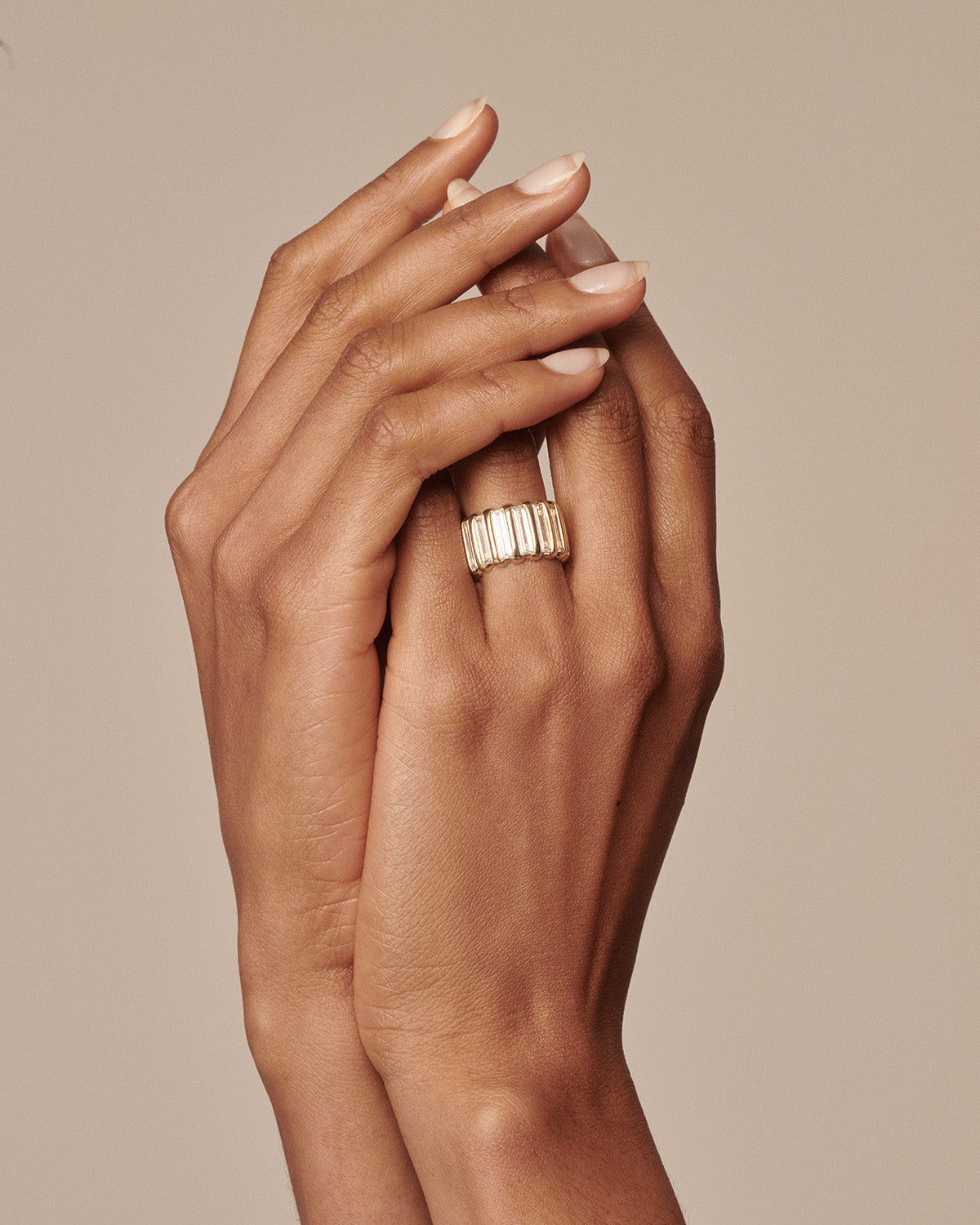 A close-up of a person's hands with manicured nails, wearing a wide gold ring with a textured ribbed design, set against a solid warm beige background. [text overlay: Francielle wears the Bold Full-Set Allure]