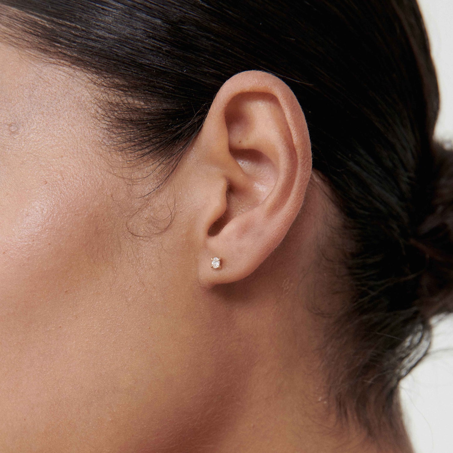 Close-up of a diamond stud earring on a woman's ear. The earring sparkles subtly against her smooth, light brown skin tone.