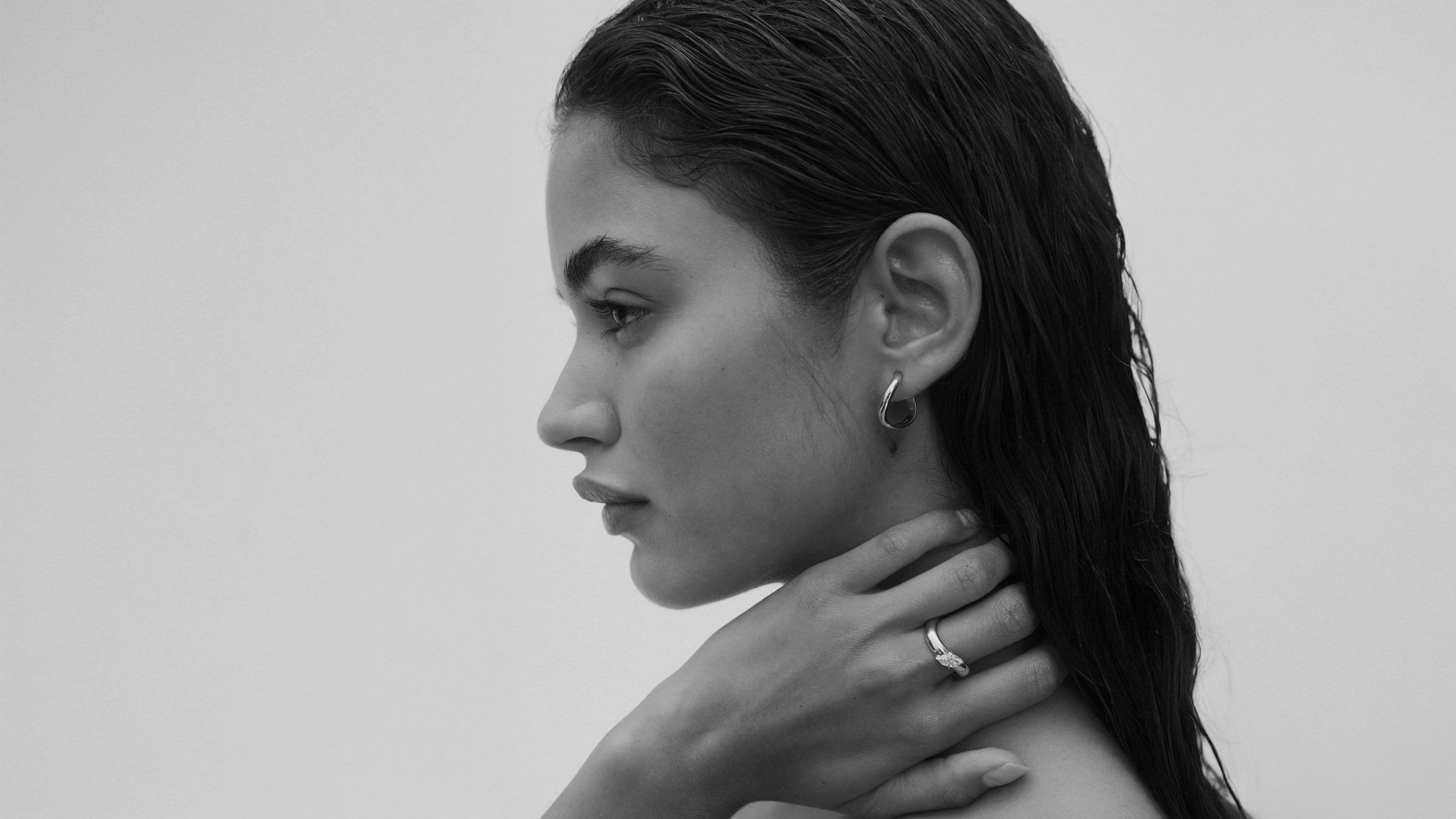 Black and white portrait of a woman with her hand on her neck against a plain background wearing Awa Hoops & Awa Wave Band with Diamond  Marquise 