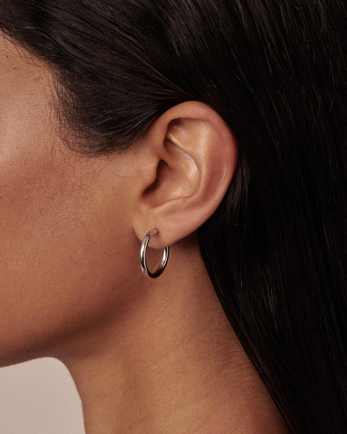 A close-up side profile of a model wearing a thin, polished silver hoop earring in her earlobe.