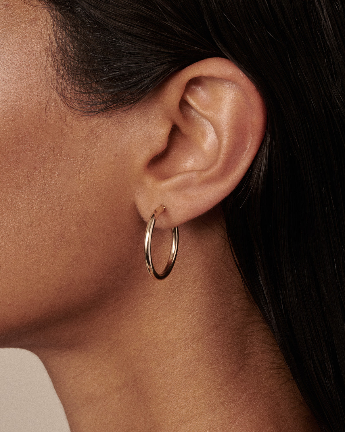 A close-up of a model's ear wearing a thin, polished gold hoop earring.