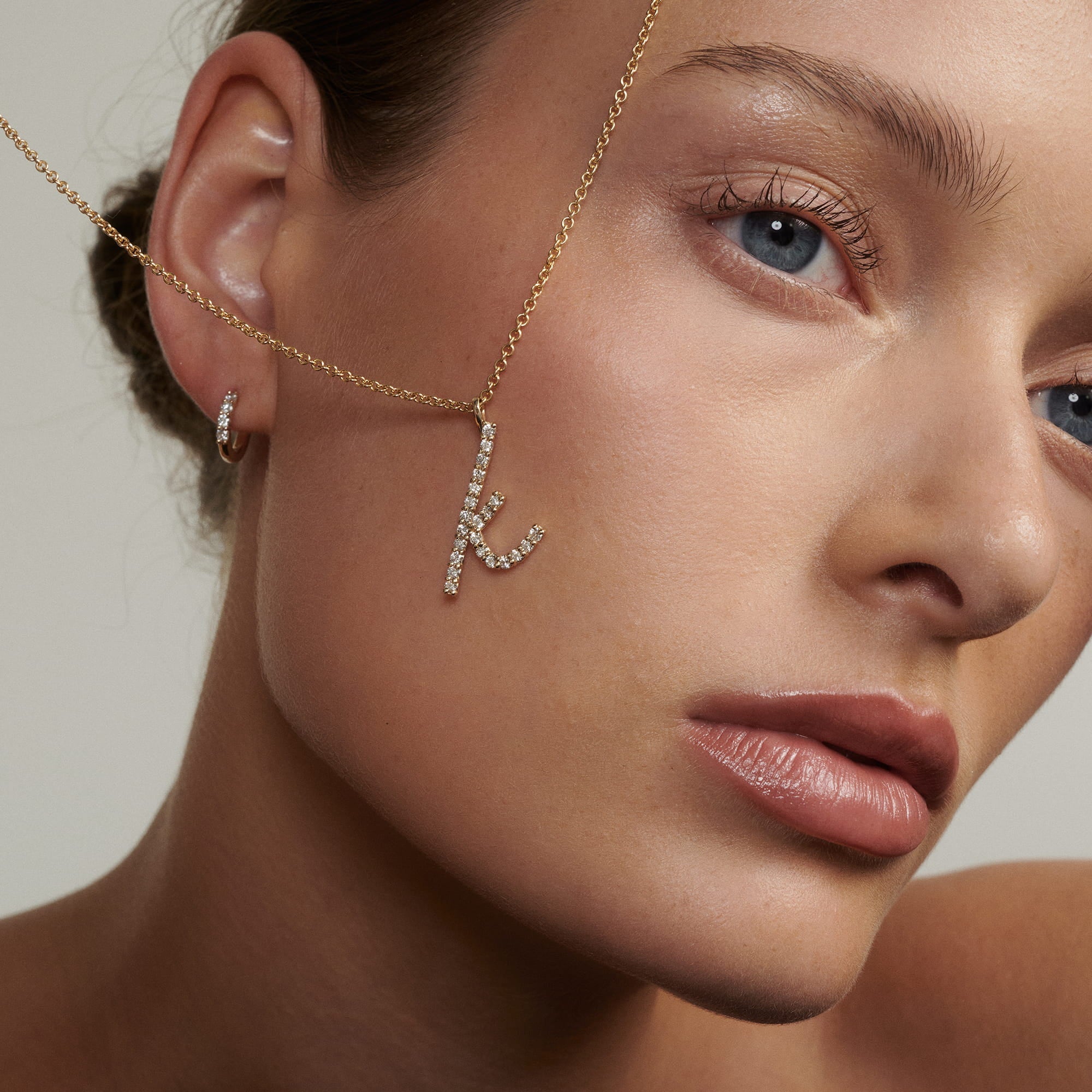 Close-up of a woman's face wearing a gold chain with a diamond letter 'K' pendant