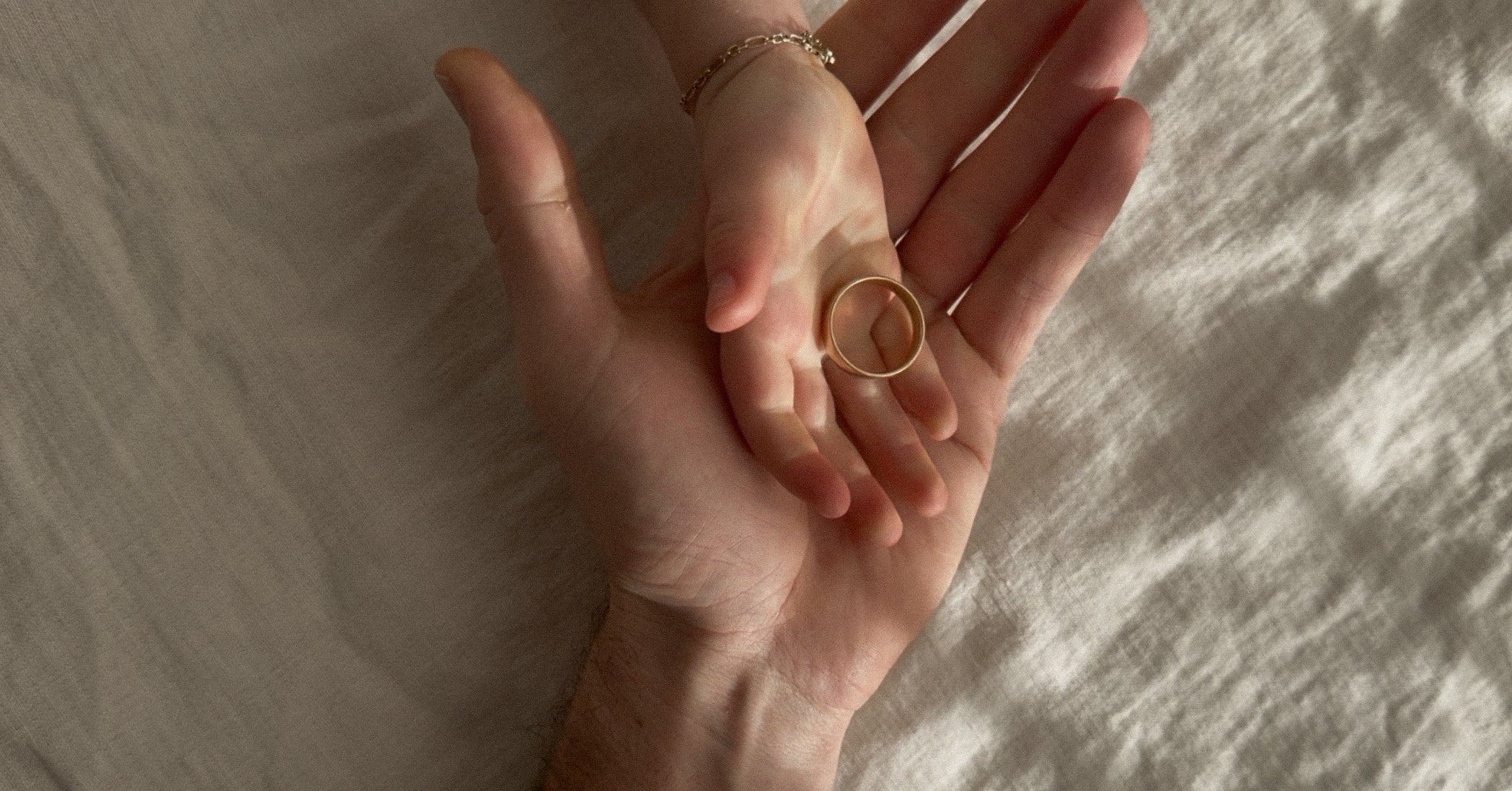 A close-up of two hands resting on a textured beige fabric surface, with a simple gold band ring held between the palms.