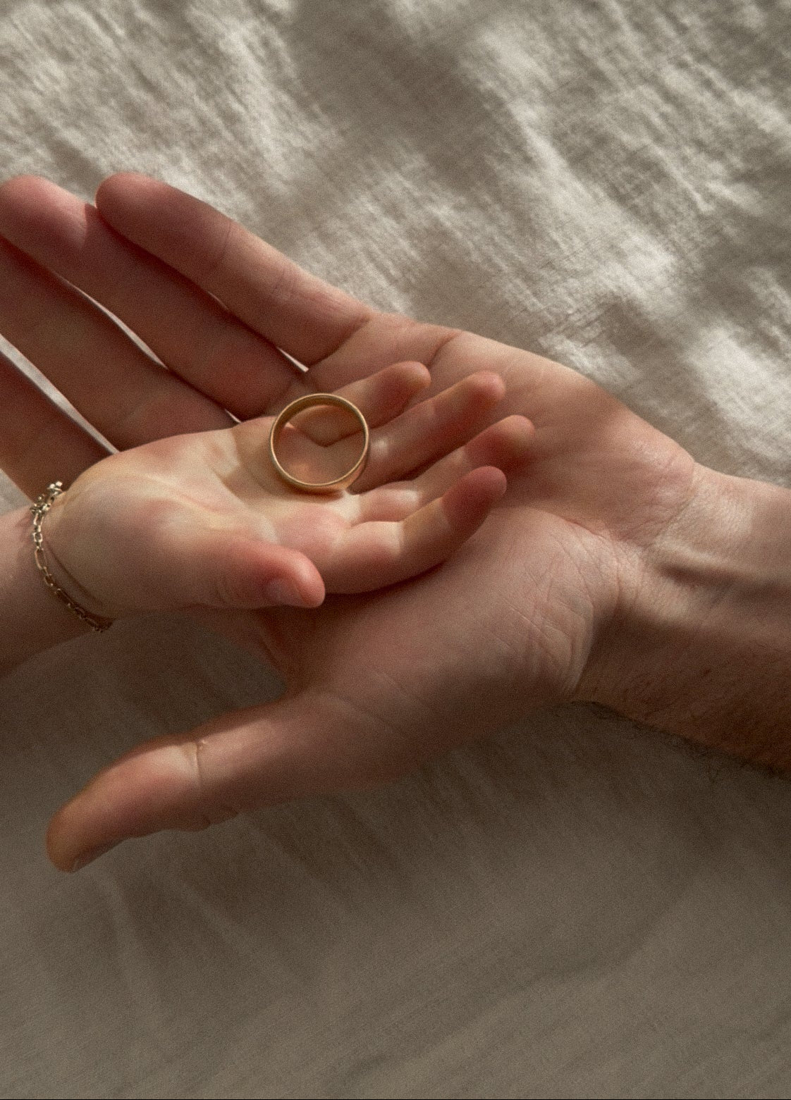 A close-up of two hands resting on a textured beige fabric surface, with a simple gold band ring held between the palms.