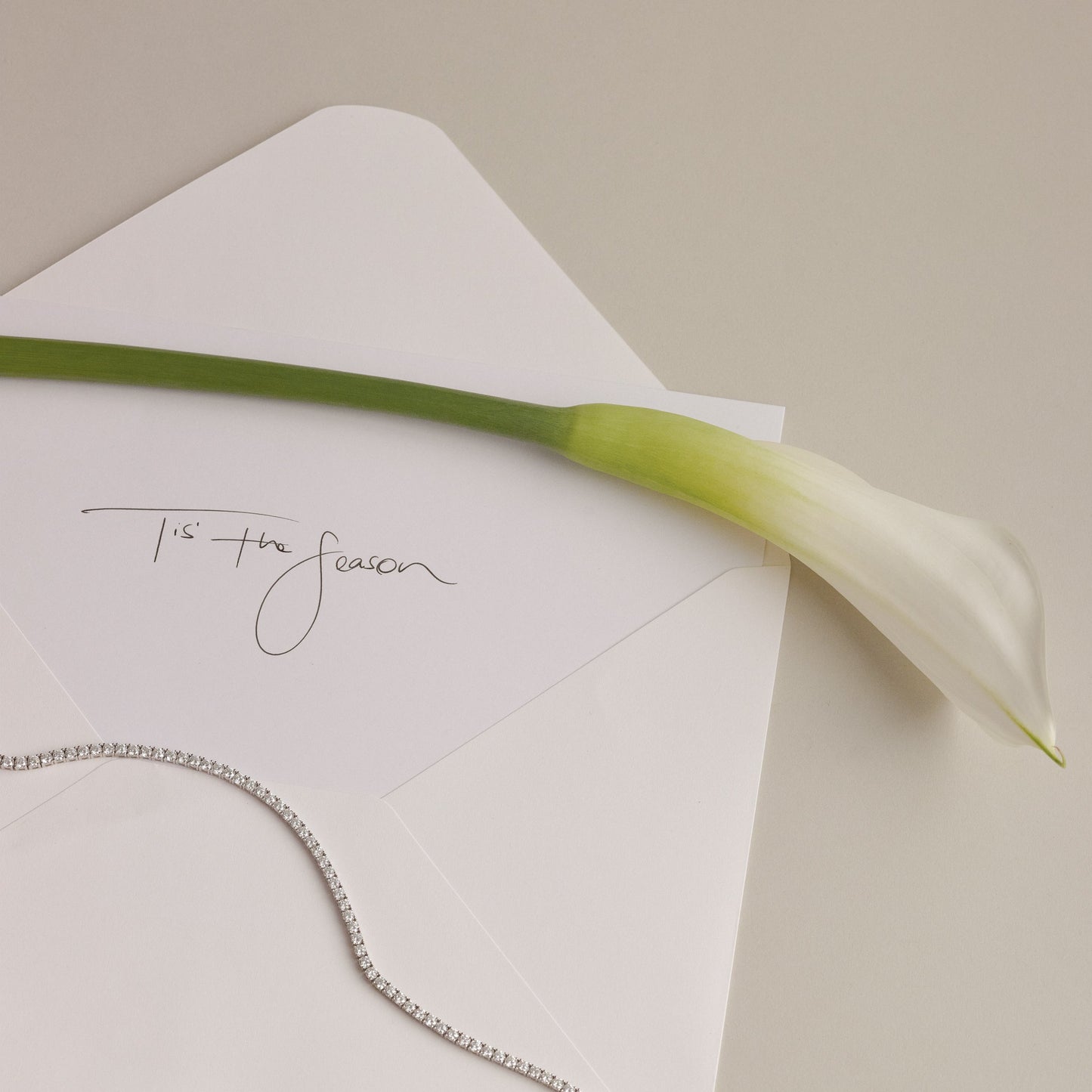 A beautiful cream envelope with a letter that writes 'Tis' the season', a tennis necklace is draped over the letter.